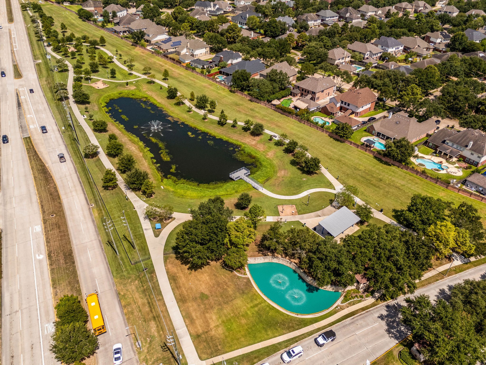 Embrace an active outdoor lifestyle with the scenic green spaces located right inside Katy Creek Ranch. This aerial view showcases the neighborhood's beautiful central park, which features a tranquil walking trail winding past a fountain-centered lake, a covered community pavilion, a dedicated playground area, and a peaceful fishing pier perfect for relaxing afternoons.