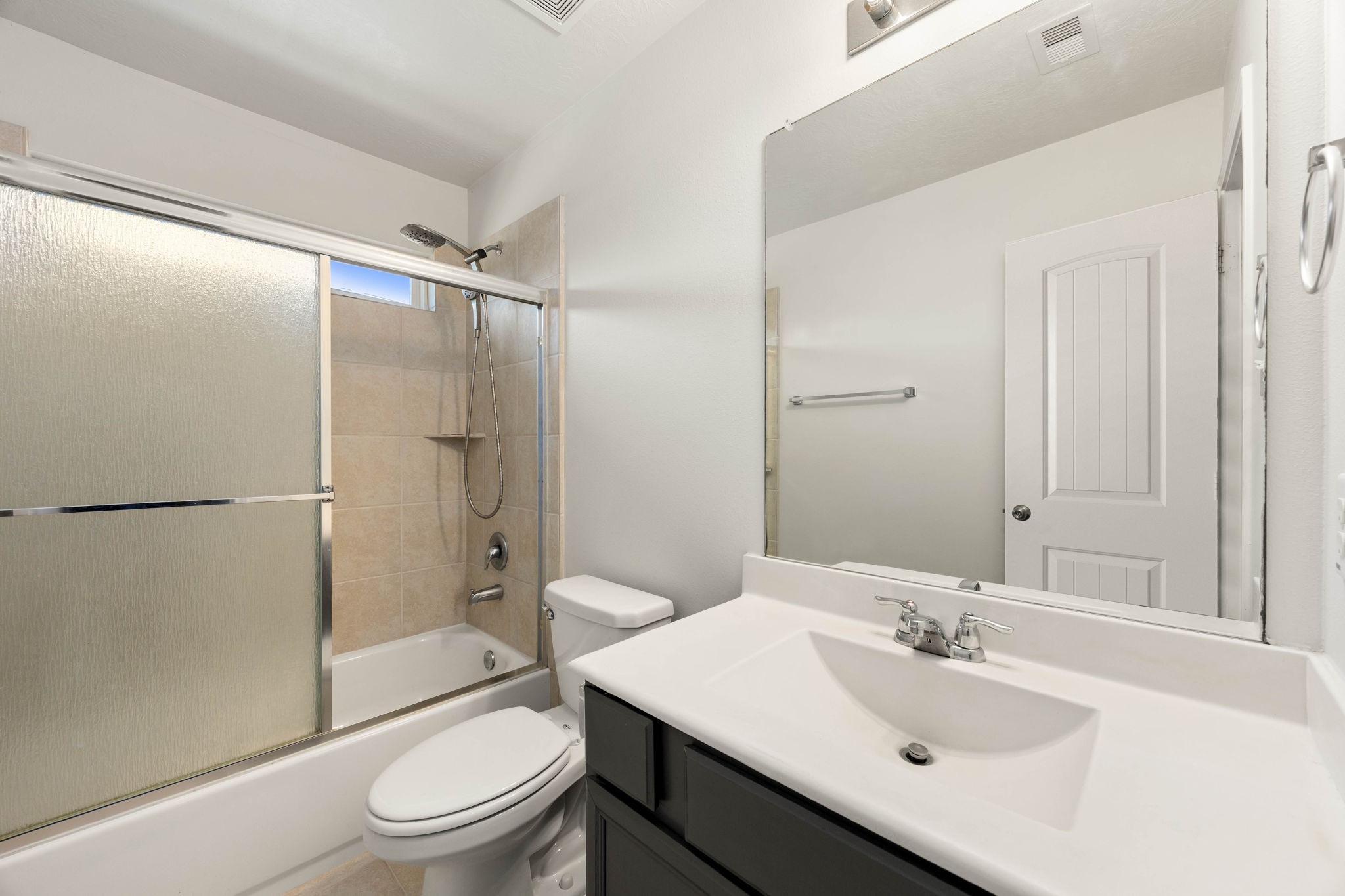 A clean and functional secondary bathroom serves the home's additional bedrooms with ease and convenience. The thoughtfully designed layout features a dark-stained vanity cabinet topped with a crisp white sink and countertop, a large seamlessly mounted vanity mirror, and a tub/shower combination complete with a neutral tile surround and frosted glass sliding enclosure doors.