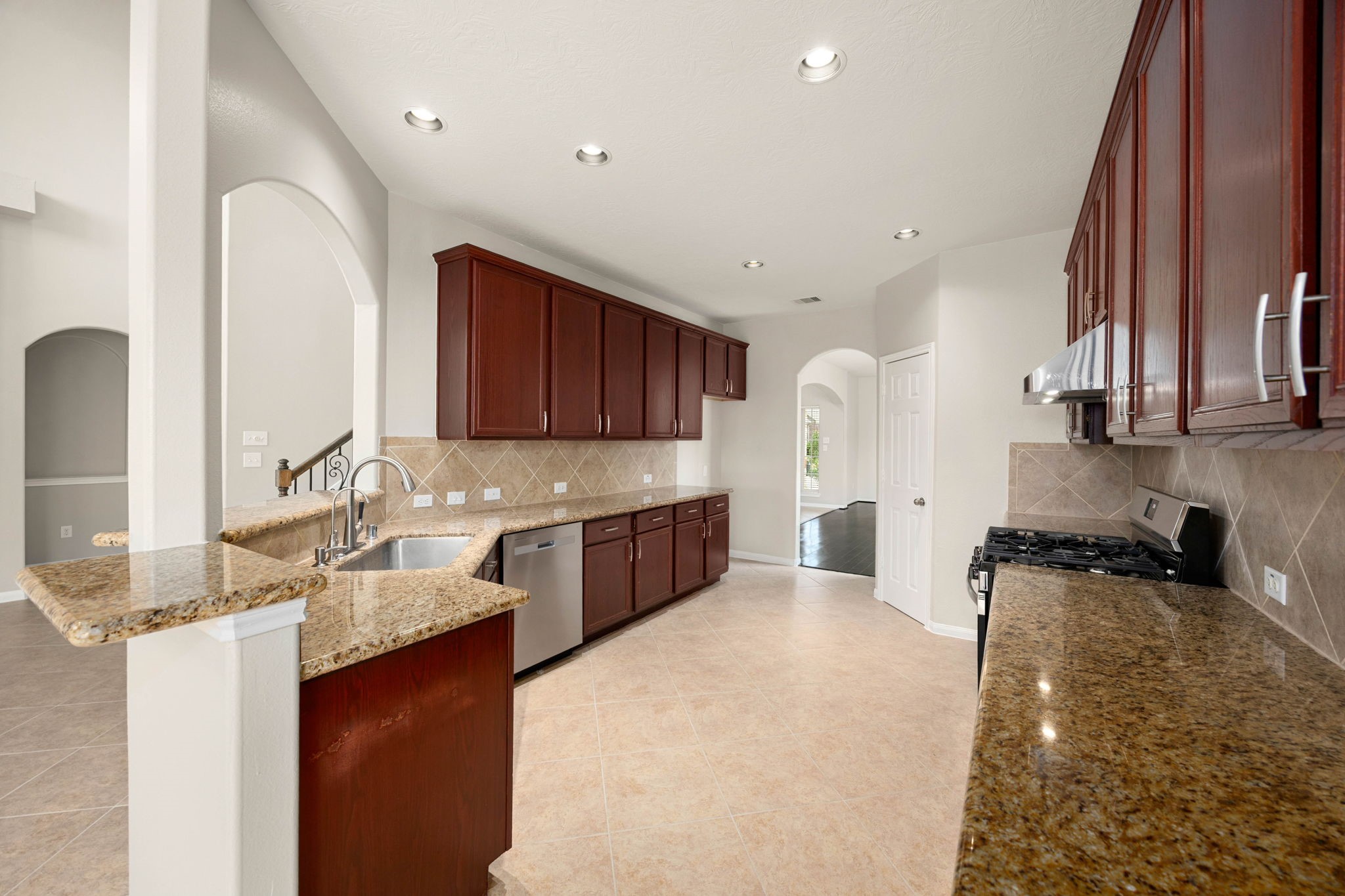Abundant granite counter space and rich, dark-stained cabinetry define this spacious kitchen, designed for maximum efficiency and storage. The functional layout features sleek stainless-steel appliances, including a under-mount double sink with a modern high-arc faucet, a gas range, and a diagonal tile backsplash, all illuminated by recessed lighting.