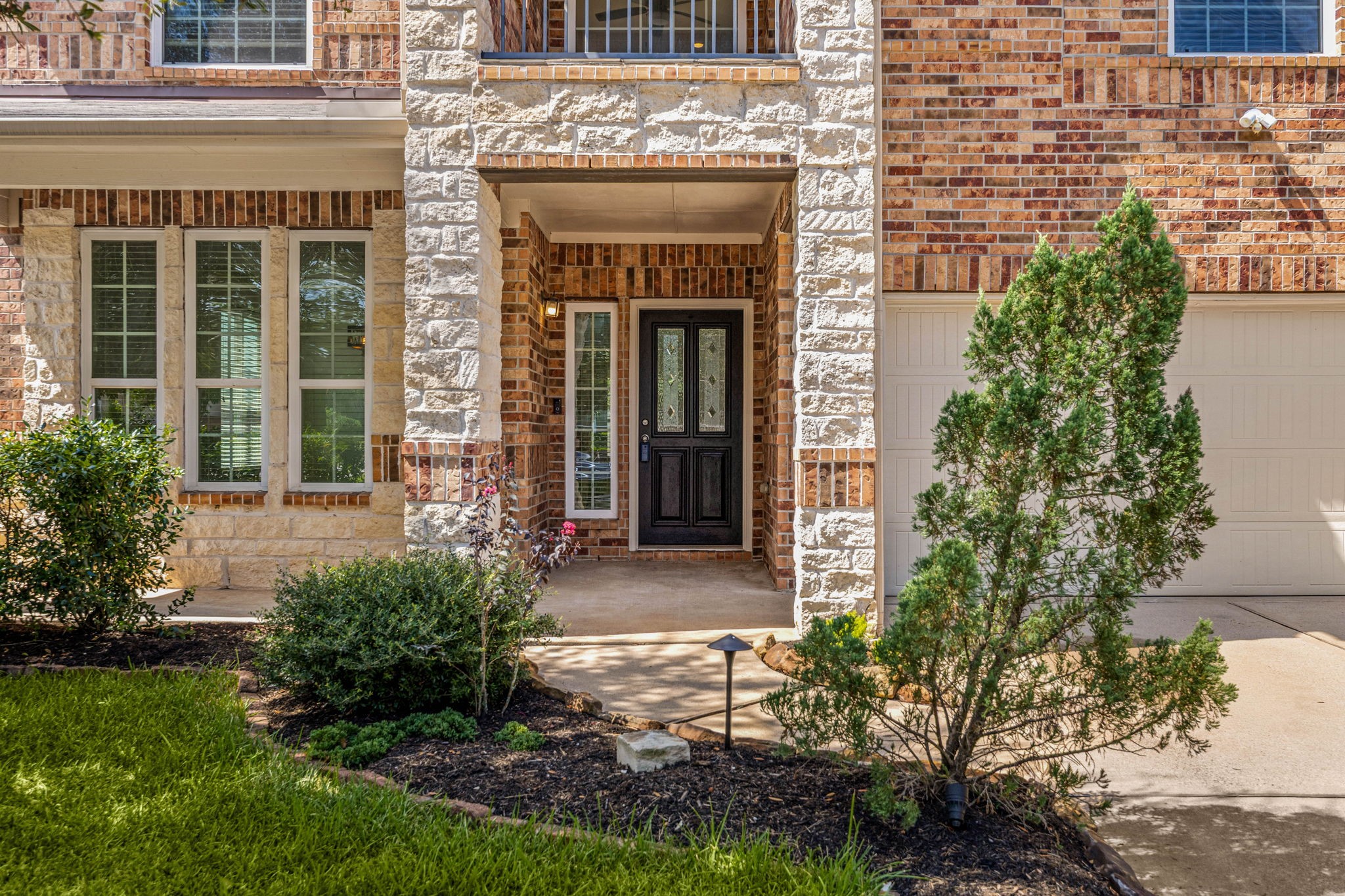 This striking front elevation perfectly combines classic brick and elegant white stone accents. The inviting, recessed front porch features a grand stone archway, manicured landscaping, and a sophisticated black entry door with decorative glass inserts, creating an undeniable sense of curb appeal the moment you arrive.
