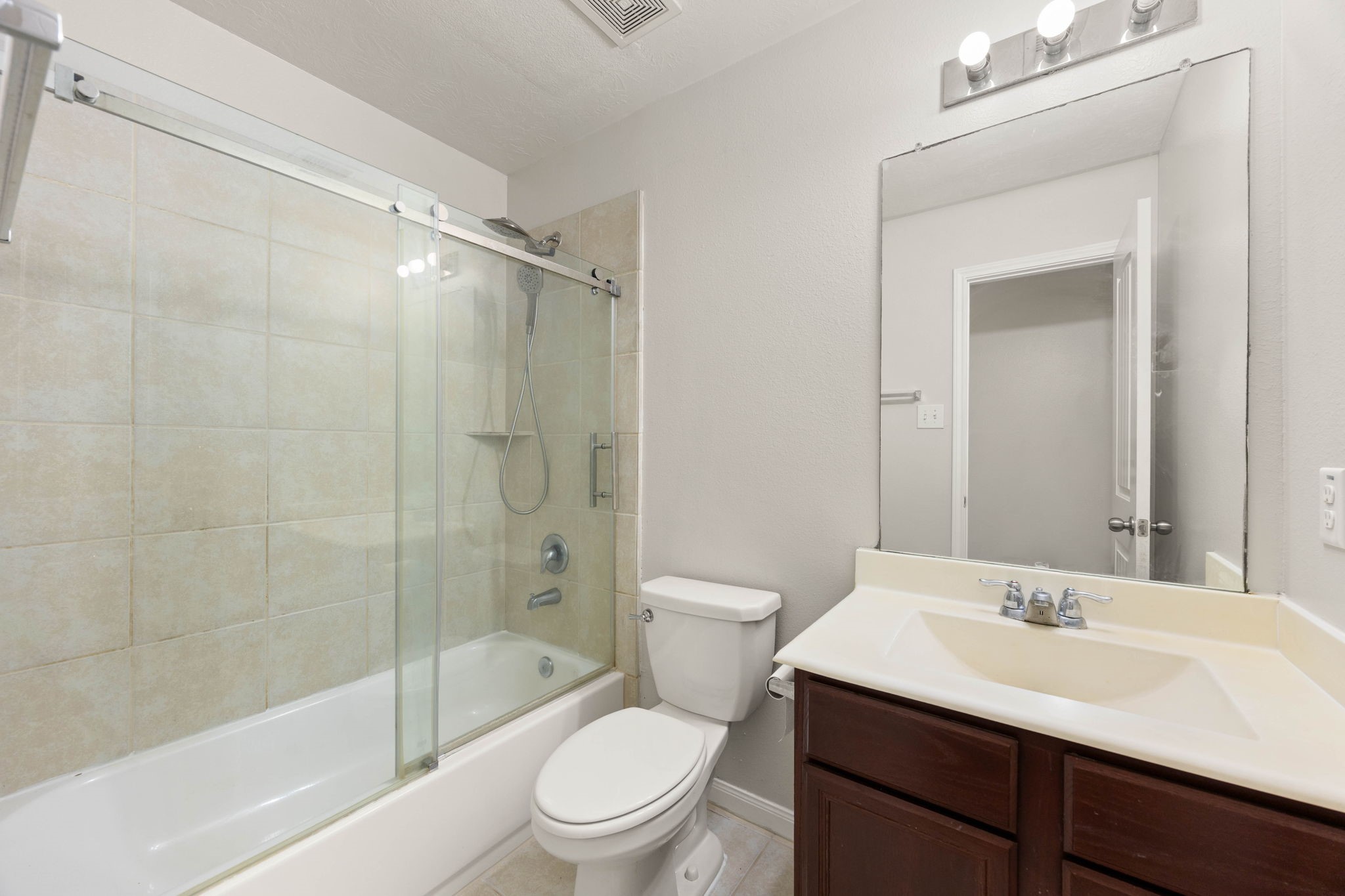 A beautifully updated secondary bathroom services the upstairs living areas, offering a perfect blend of style and function. The space features a dark wood vanity with a crisp white countertop, a large vanity mirror with modern lighting, and a tub/shower combination upgraded with an elegant, frameless sliding glass door layout and a matching tile surround.