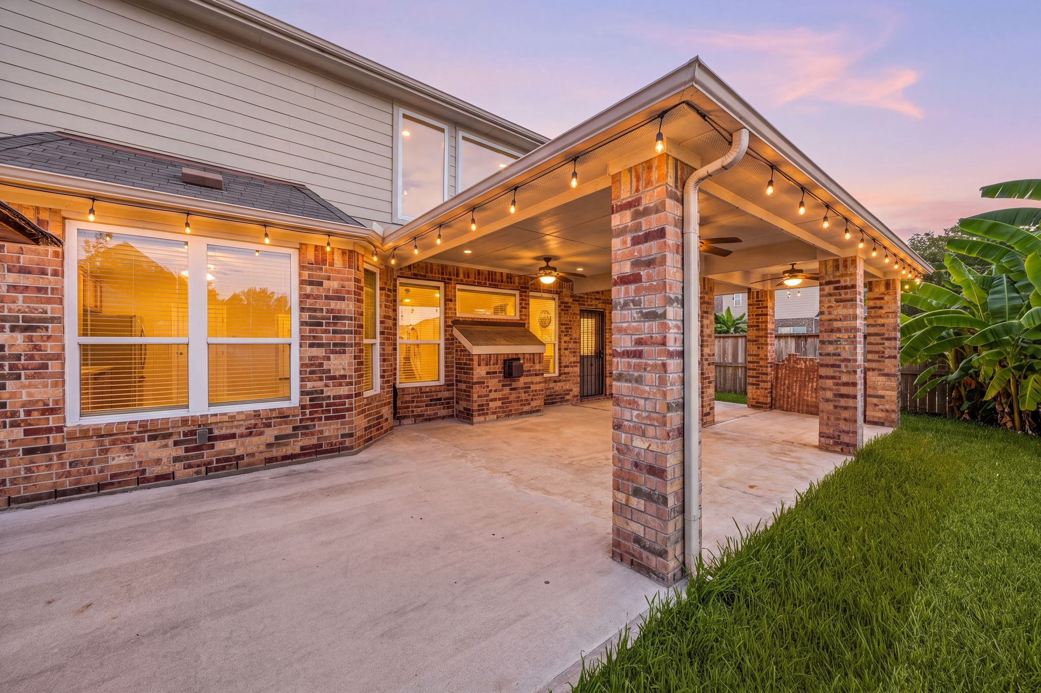 An expansive covered patio addition transforms this backyard into an outdoor entertainer's paradise, highlighted by robust brick pillars. The massive concrete extension features multiple integrated ceiling fans, a functional built-in service pass-through structure, and a deep overhang that provides continuous shade and comfortable outdoor living space year-round.