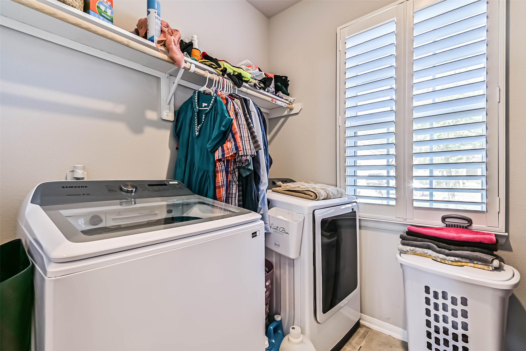 Dedicated laundry room featuring washer and dryer space, overhead shelving, hanging storage, and a window with plantation shutters.