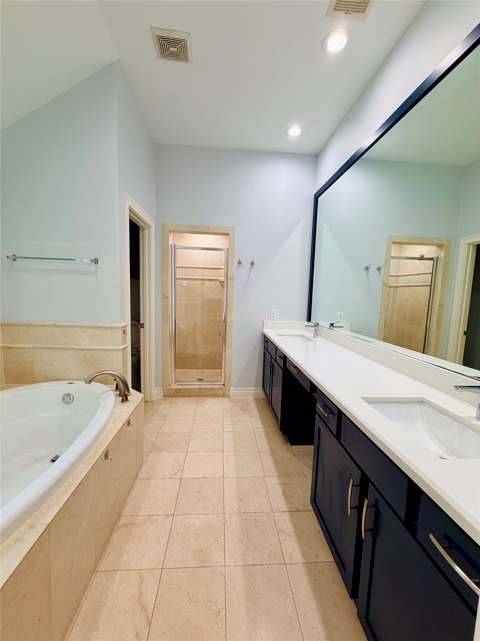 This bright and spacious primary bathroom has recessed lighting, tile flooring, dual-vanity, tub, and a large walk-in shower with tile surround and glass enclosure, and a thoughtfully designed layout.