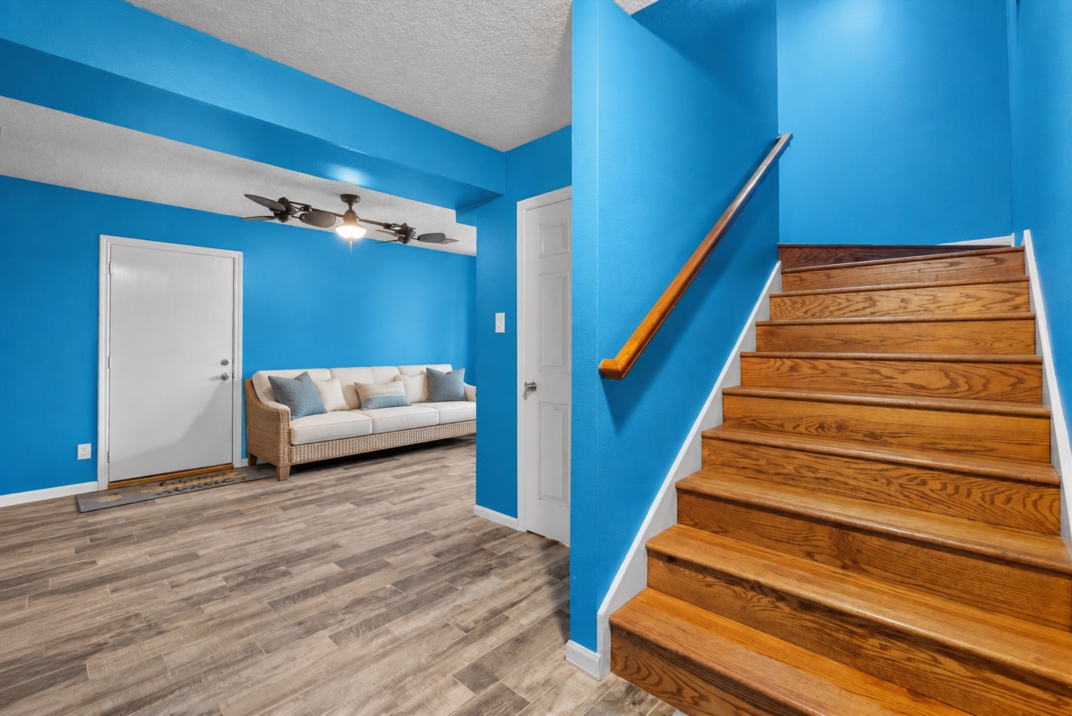 A BRIGHT ENTRYWAY FEATURES WOOD-LOOK TILE FLOORING, VIBRANT COASTAL BLUE WALLS, AND A WARM WOOD STAIRCASE. THE OPEN LAYOUT CONNECTS TO A VERSATILE SITTING AREA WITH EASY ACCESS TO THE ADJACENT ROOMS.