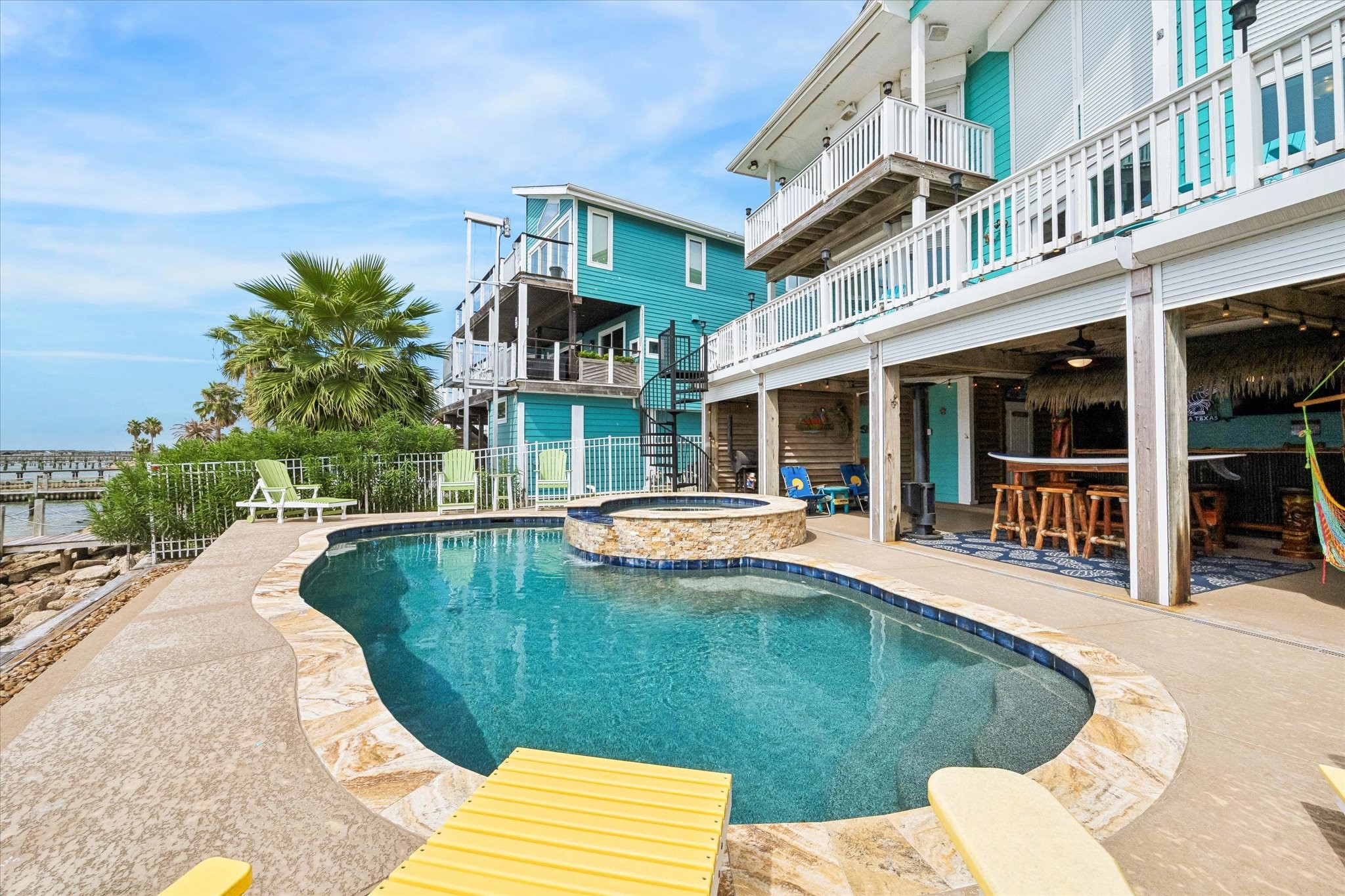 THE REAR EXTERIOR FEATURES MULTIPLE BALCONIES, A SPIRAL STAIRCASE, A COVERED PATIO, AND A WATERFRONT POOL AND SPA.