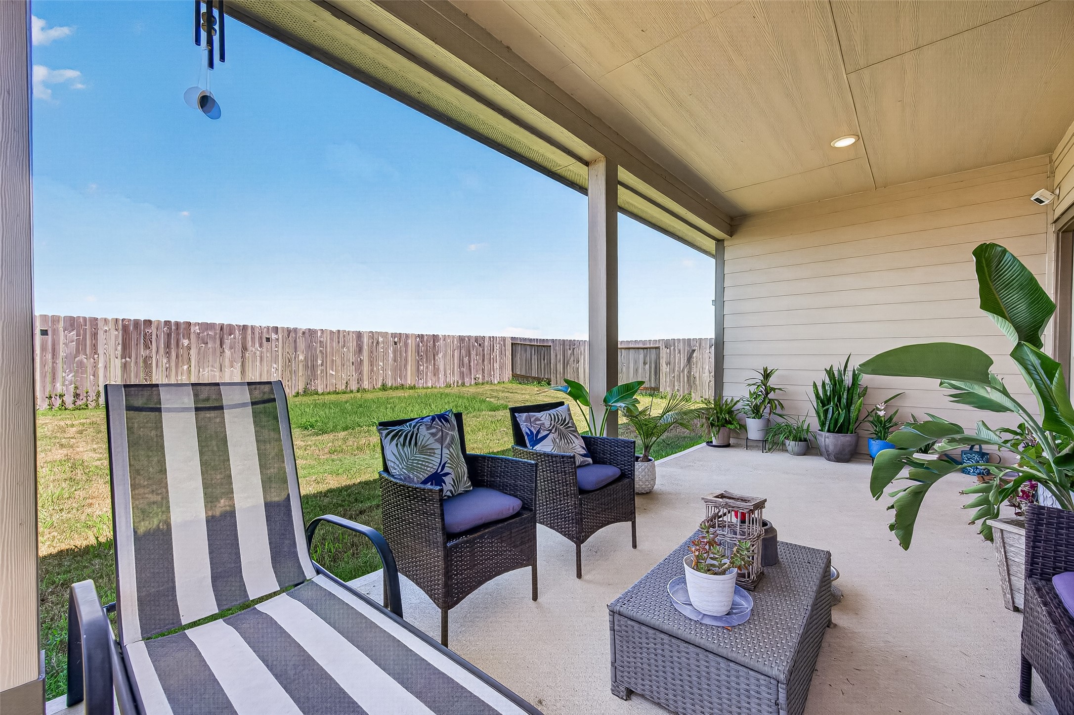 Relax in this inviting covered patio featuring comfortable seating, lush greenery, and a spacious fenced yard perfect for outdoor enjoyment.