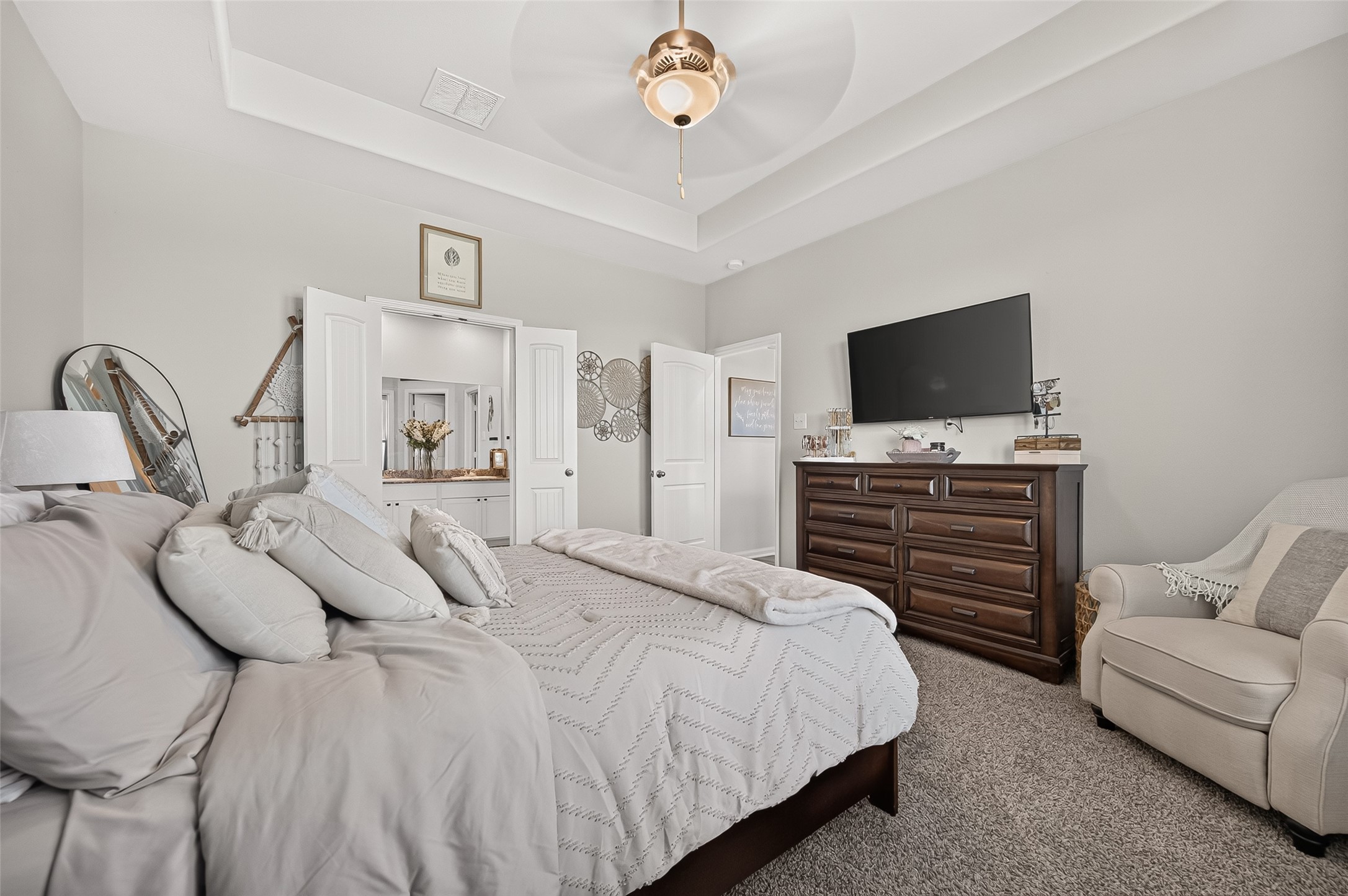 Spacious bedroom featuring a cozy bed with plush pillows, a large dresser with a mounted TV, and an inviting armchair, all set under a stylish ceiling fan and soft neutral tones.