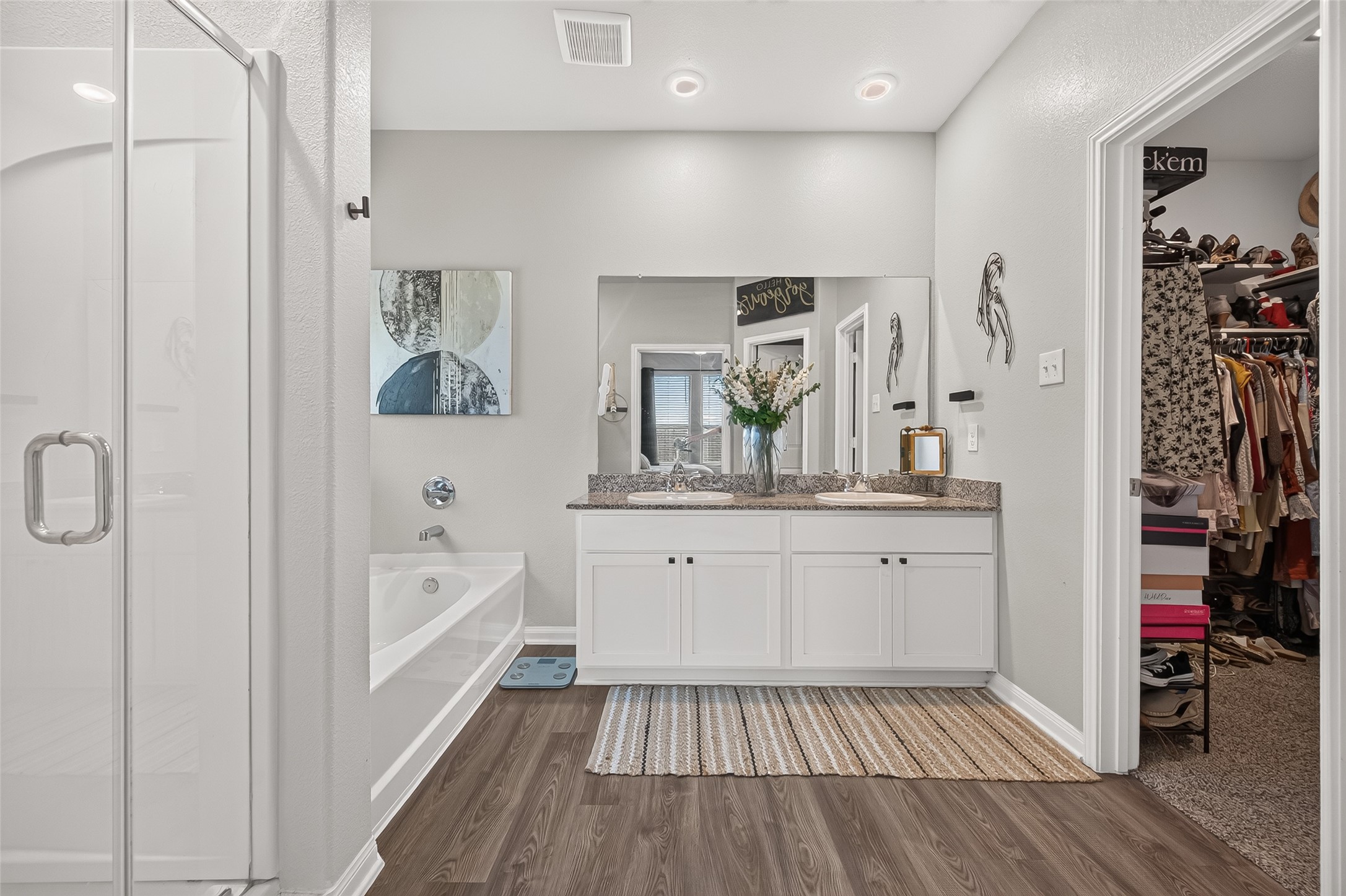 Spacious bathroom featuring a double vanity with granite countertops, a soaking tub, and a glass-enclosed shower, complemented by a walk-in closet.
