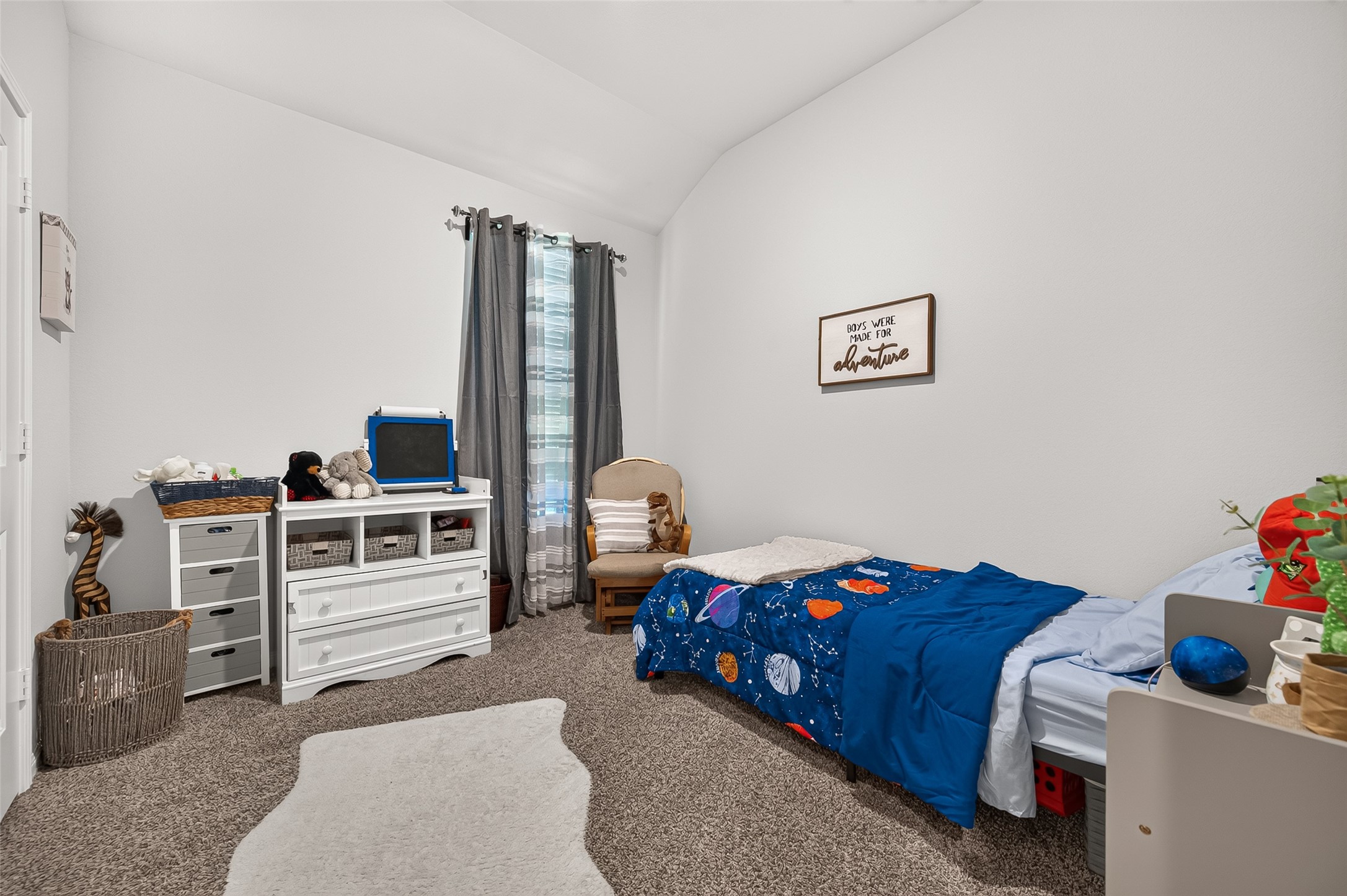 Cozy children's bedroom featuring plush carpeting, a cheerful space-themed bedspread, and ample storage with a white dresser and shelves. Natural light streams in through the window, creating a bright and inviting atmosphere.