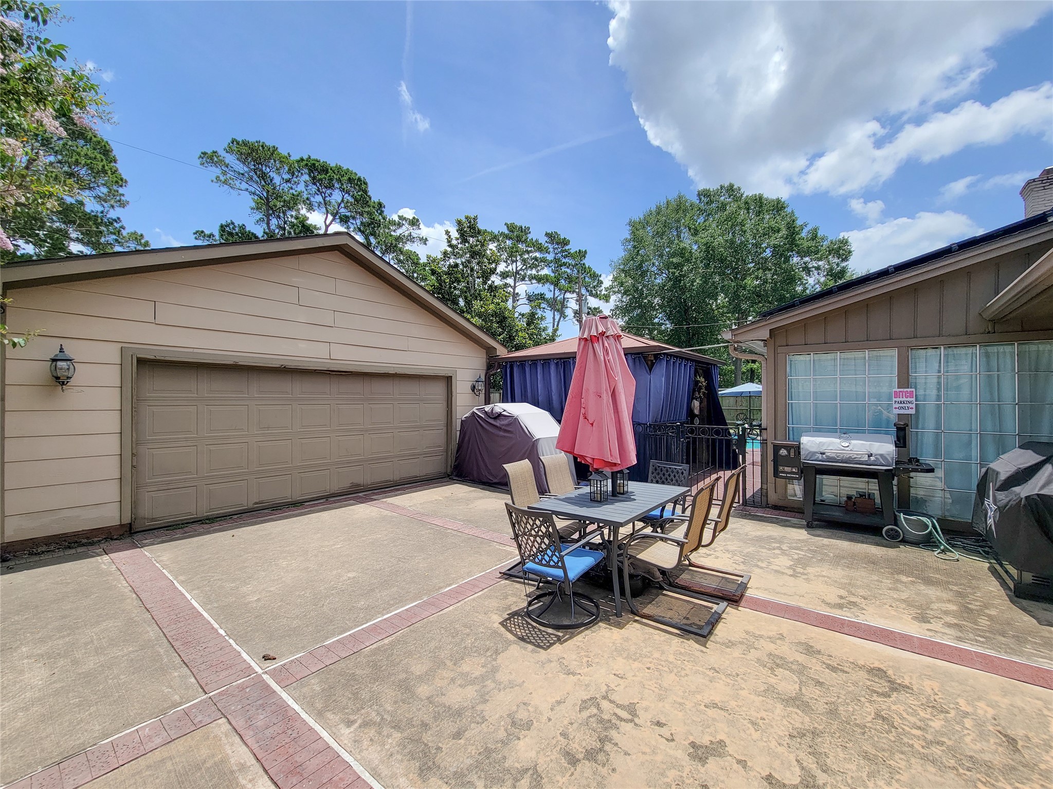 A spacious backyard patio area with a dining set and umbrella, a grill, and ample space for outdoor activities. It is bordered by a garage and features greenery in the background, offering a private and inviting space for entertaining.