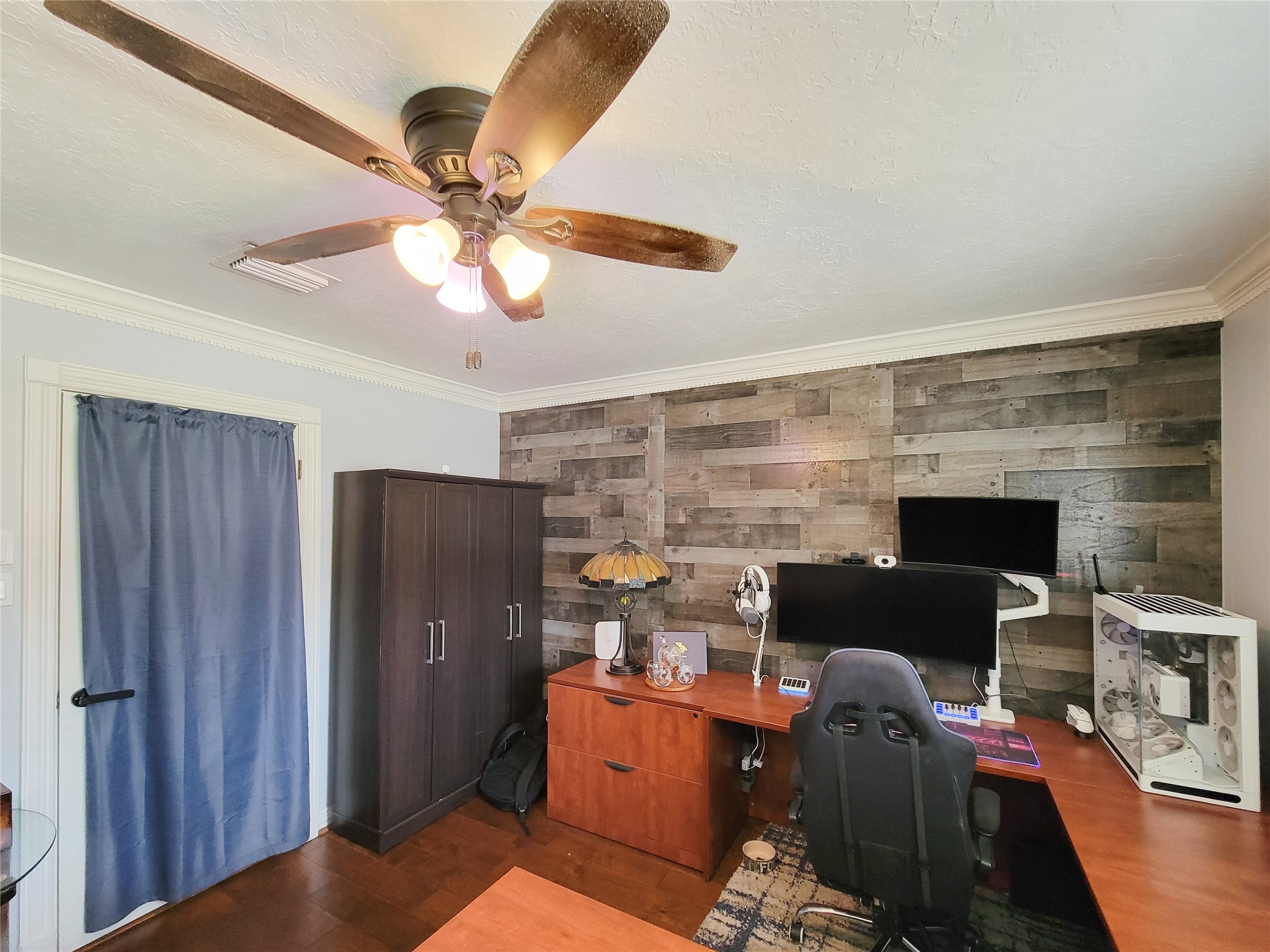 A cozy home office featuring a modern ceiling fan, wooden accent wall, and ample workspace with a large desk and shelving. The room includes a comfortable office chair, built-in computer setup, and stylish lighting, creating an inviting and functional work environment.