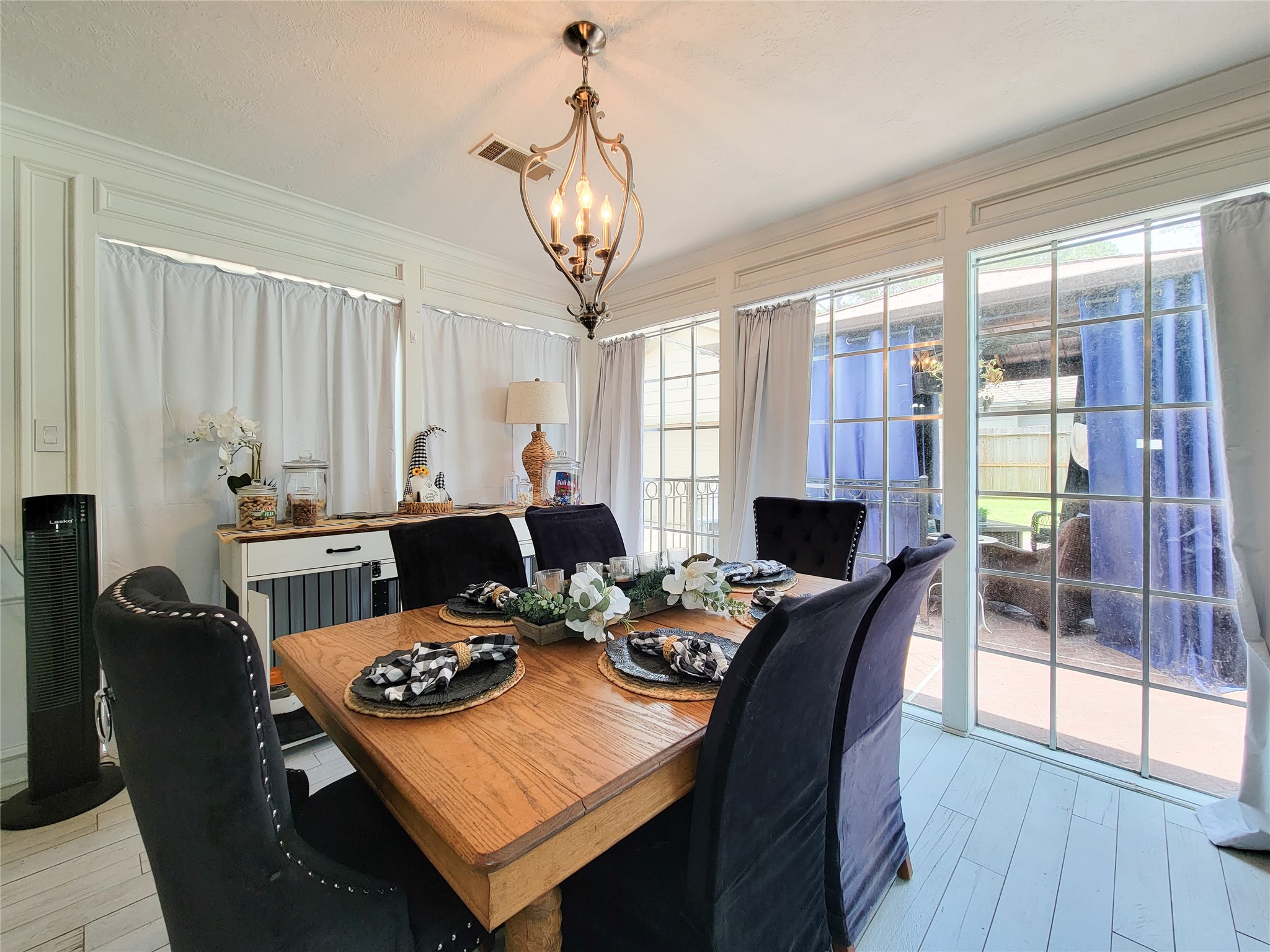 Bright dining area with large windows, elegant chandelier, and a wooden table set for six. The space features light wood flooring and leads to an outdoor patio area, creating an inviting atmosphere for gatherings.