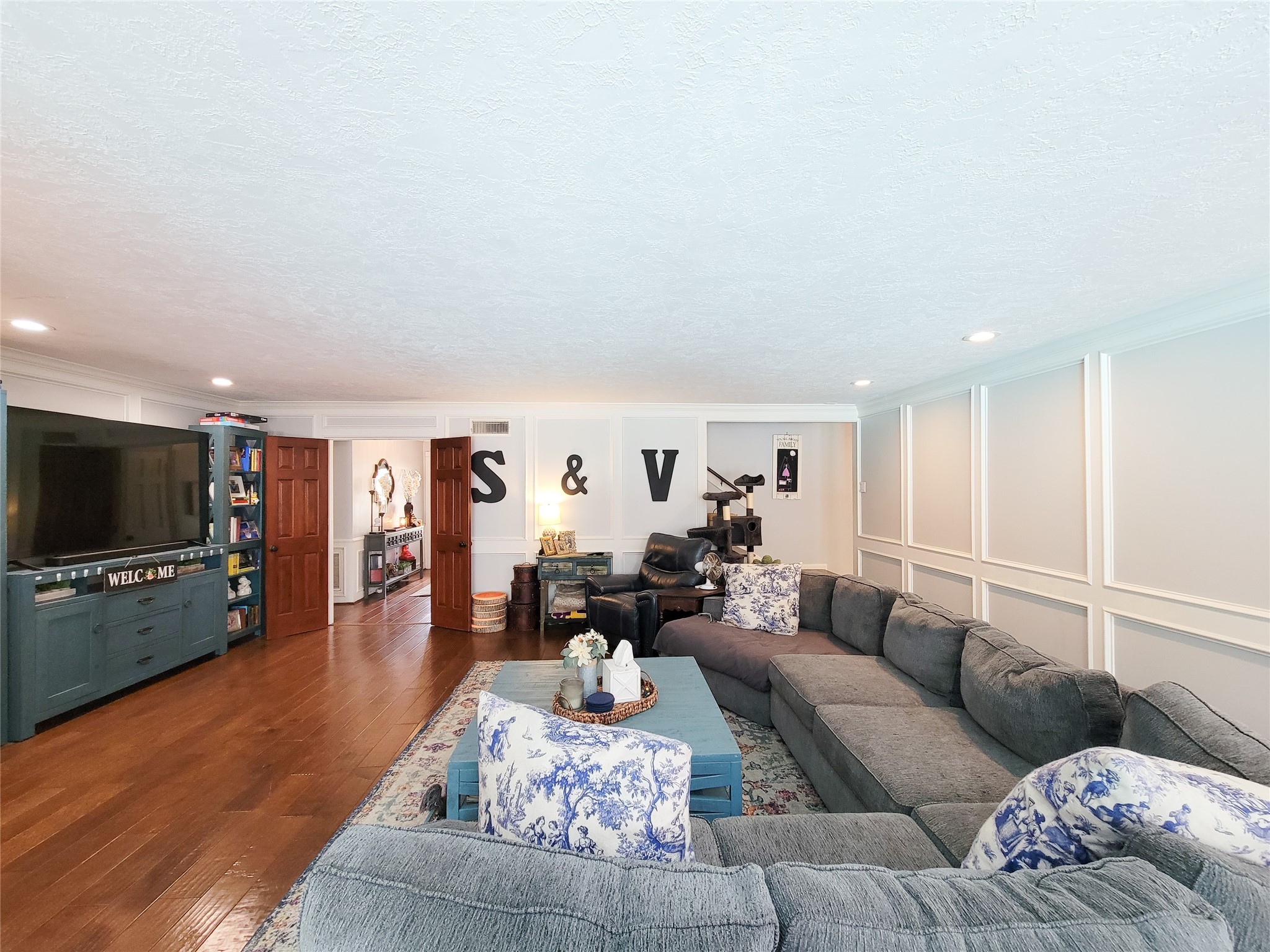 Spacious living room with elegant wood flooring, a large sectional sofa, and ample natural light. Features a cozy TV area, decorative wall paneling, and open entryways leading to adjacent rooms.