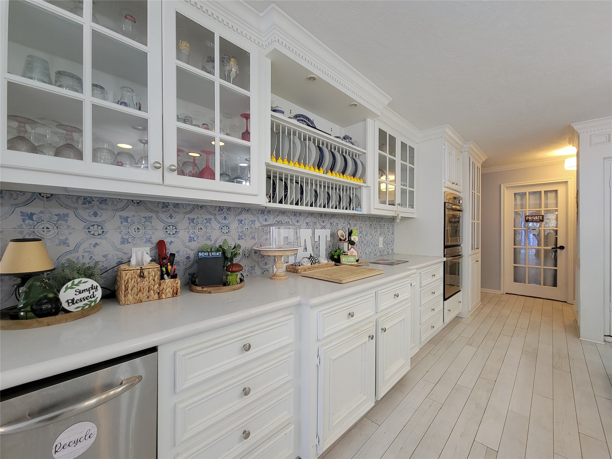 This kitchen features elegant white cabinetry with glass doors, a decorative blue and white tile backsplash, and ample counter space. It includes a built-in oven and a dishwasher, with a bright, inviting atmosphere enhanced by light wood flooring and recessed lighting.