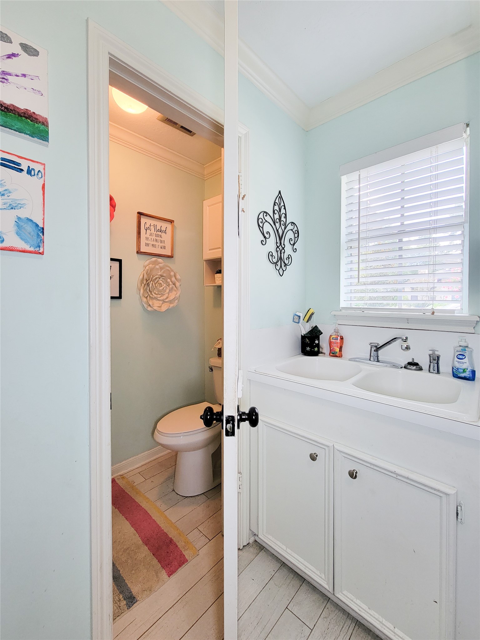 This photo showcases a compact bathroom with pastel blue walls, featuring a toilet, a white sink with cabinets, and a window with blinds. Decorative elements include wall art and a small rug, creating a cozy, inviting space.
