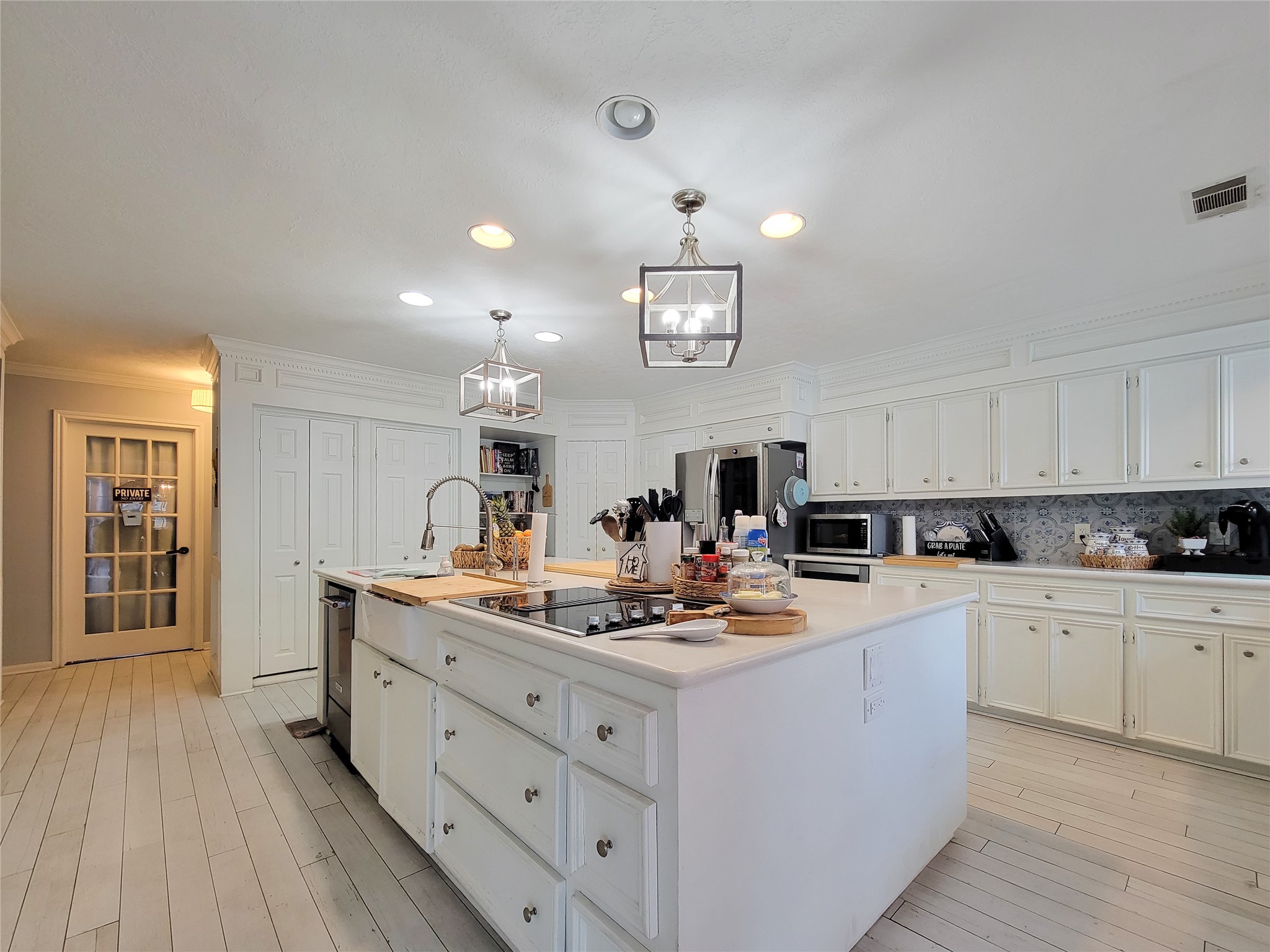 This spacious kitchen features modern lighting, ample white cabinetry, and a large island with a cooktop and sink. The light wood flooring and stainless steel appliances add a contemporary touch, while a glass-paneled door leads to additional storage space.