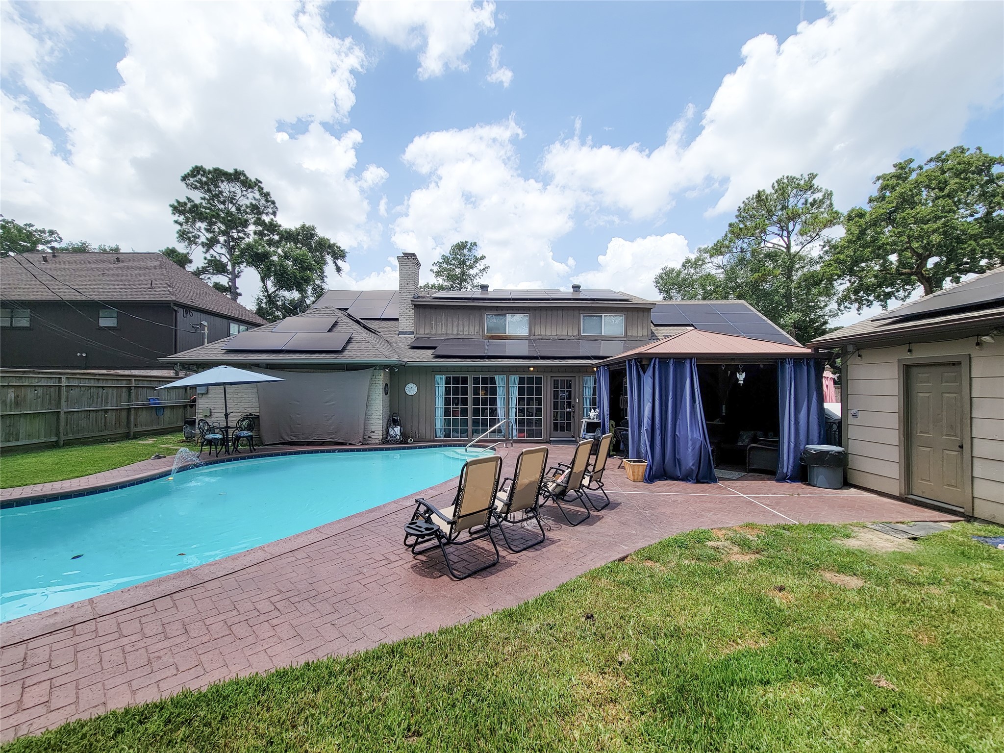 This photo features a spacious backyard with a large swimming pool, surrounded by a brick patio. There's ample seating with several reclining chairs, a covered outdoor area with curtains, and mature trees providing natural shade. The house has solar panels and a fence for privacy, offering a perfect blend of relaxation and functionality.