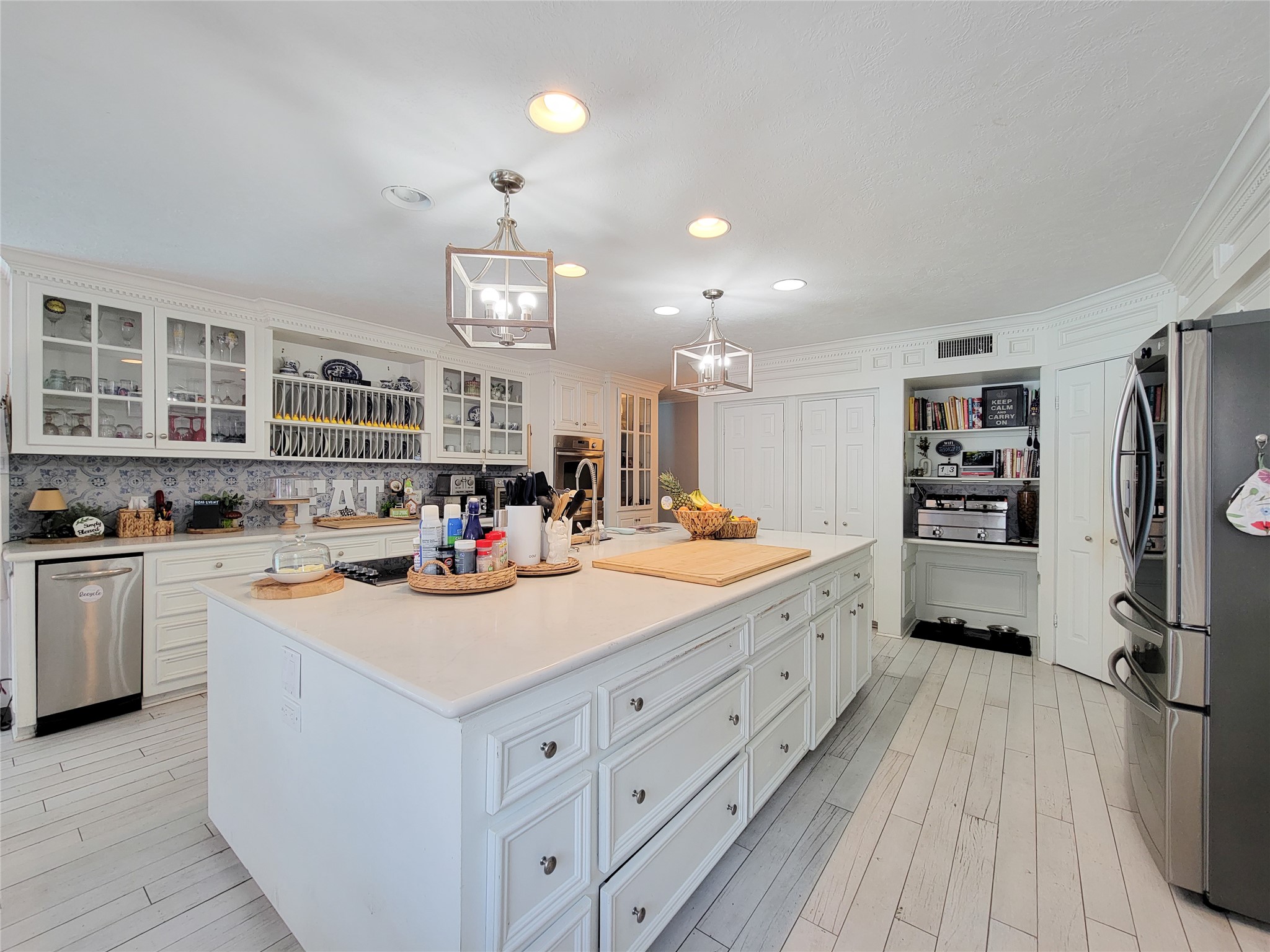 This spacious kitchen features a large island with ample storage, modern stainless steel appliances, and elegant white cabinetry with glass fronts. The space is bright and inviting, enhanced by stylish pendant lighting and a sleek, open layout perfect for cooking and entertaining.