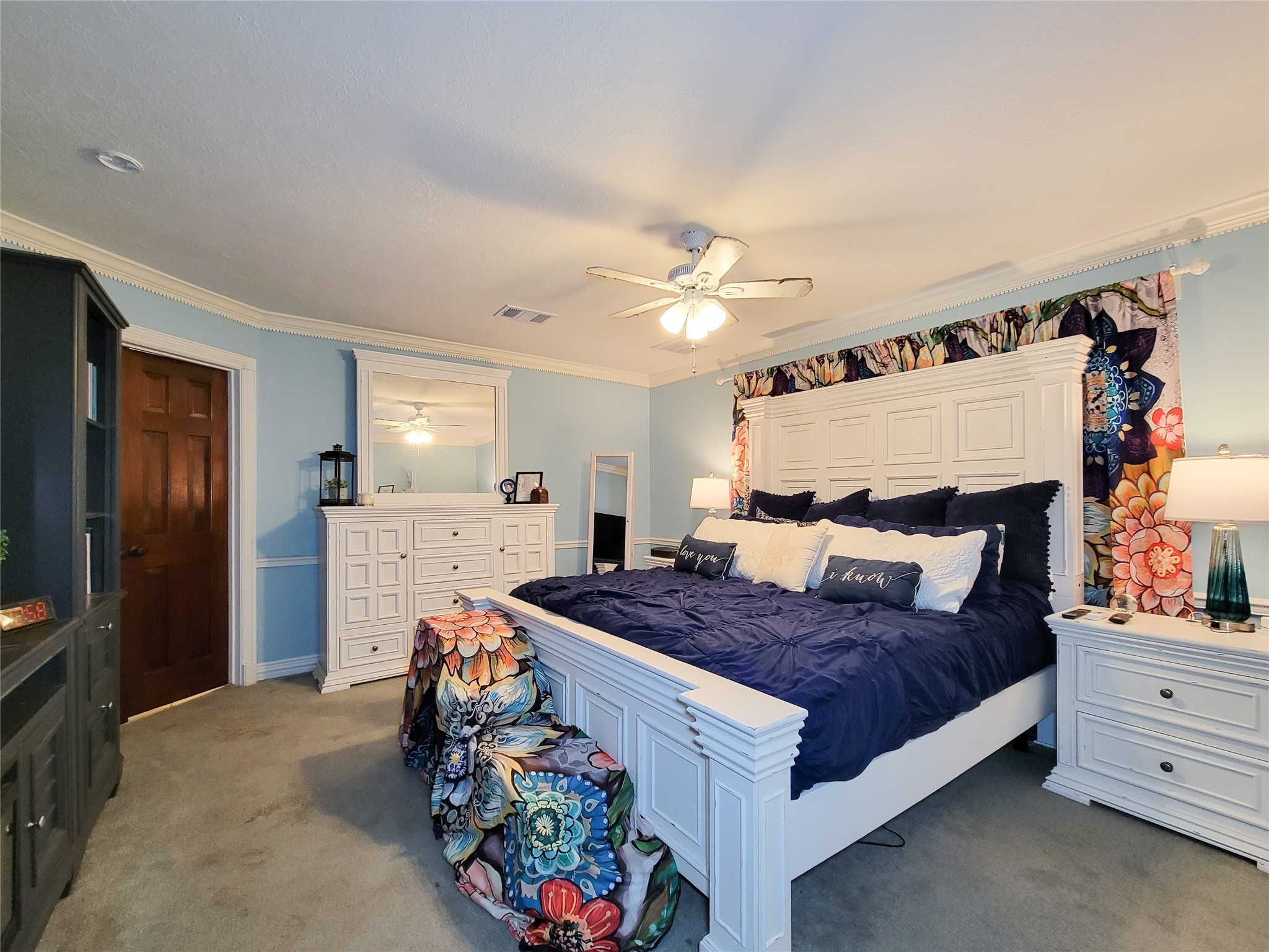 This spacious Primary bedroom features a ceiling fan, soft lighting, and colorful floral accents, creating a cozy and inviting atmosphere. This spacious Primary bedroom features a ceiling fan, soft lighting, and colorful floral accents, creating a cozy and inviting atmosphere.