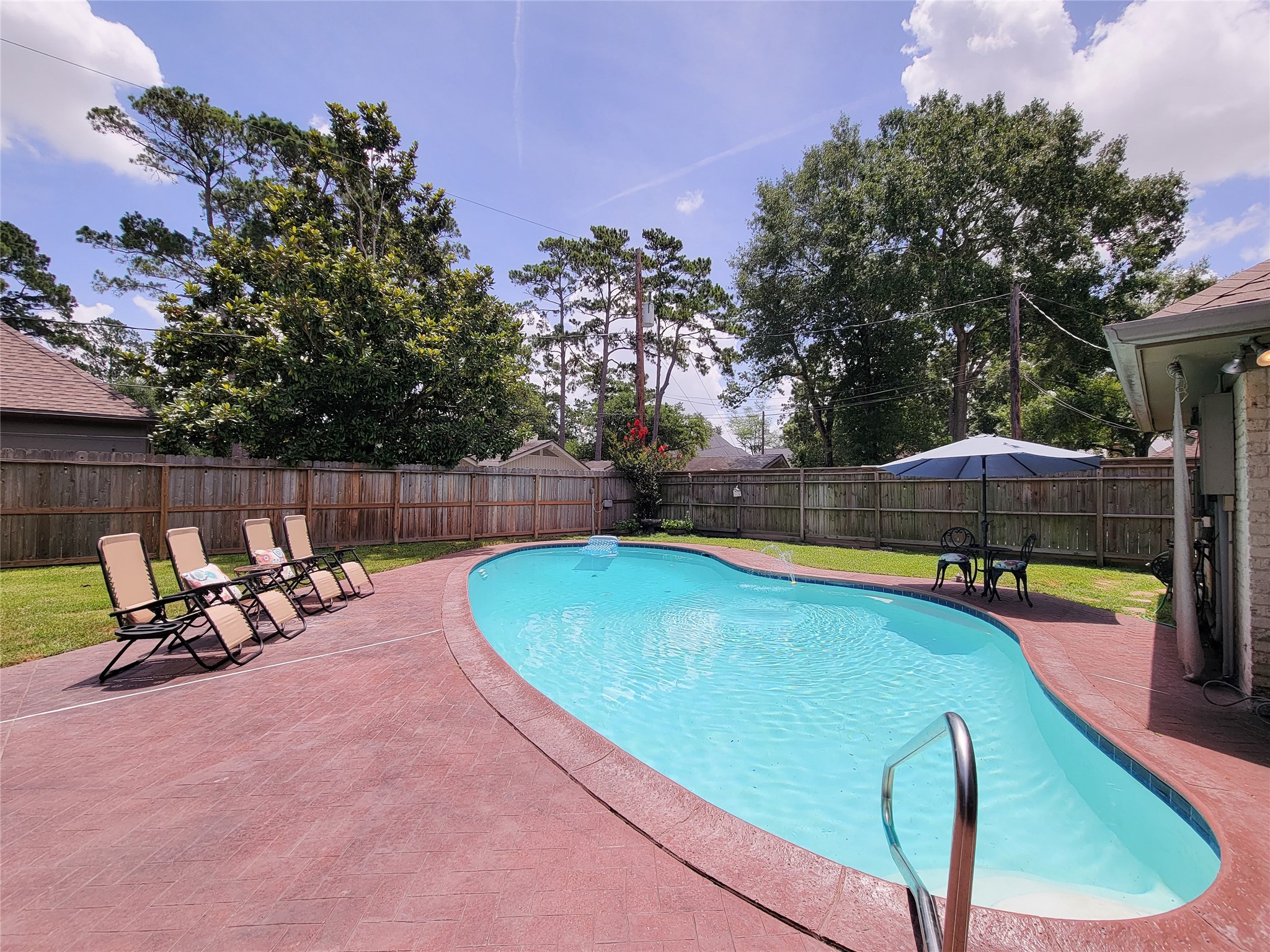 This photo showcases a spacious backyard with a curvy pool, surrounded by a well-maintained patio. There are several lounge chairs and a seating area with an umbrella, perfect for relaxation and entertaining against a backdrop of mature trees and a wooden fence.