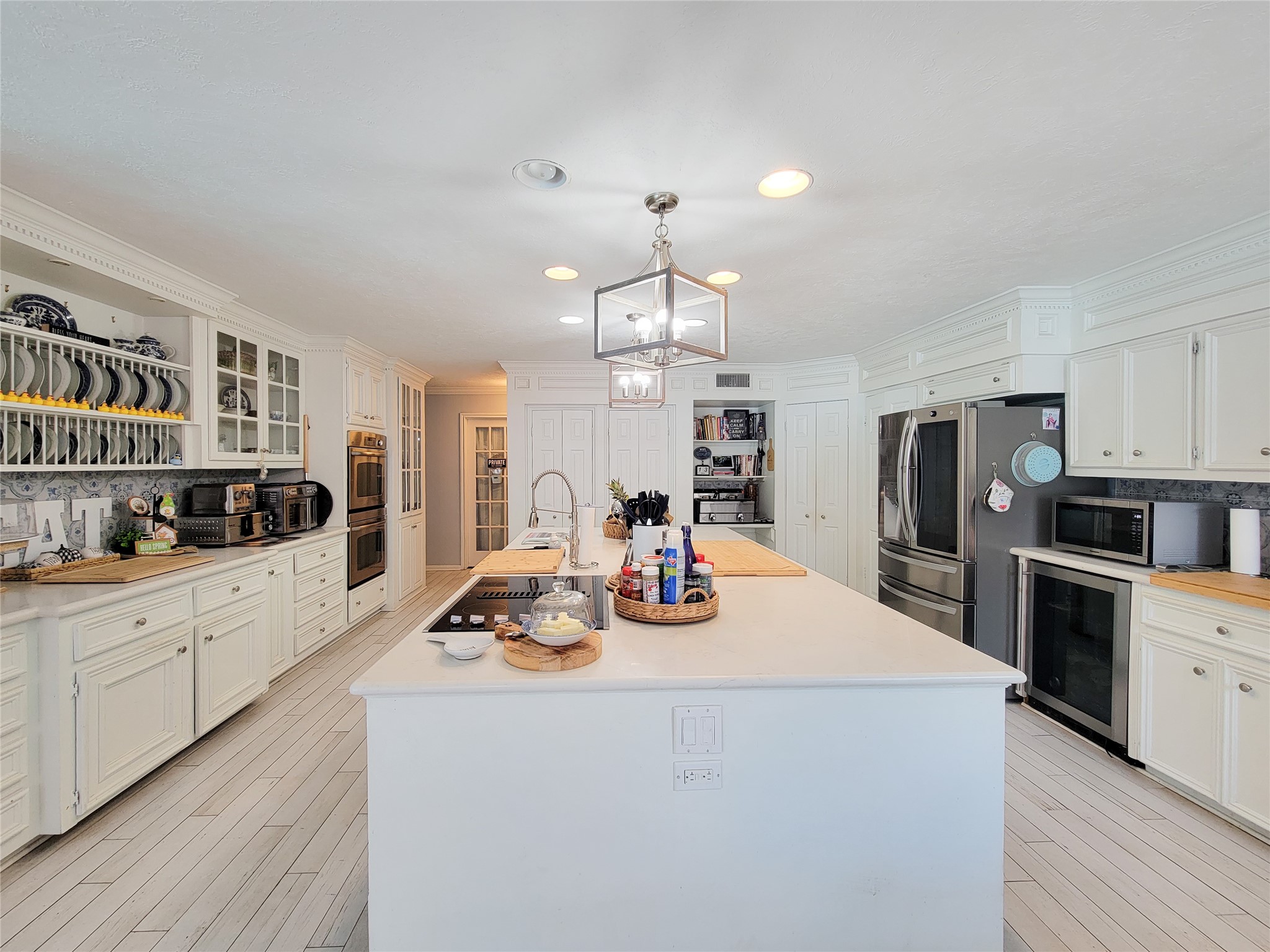 This spacious kitchen features white cabinetry, modern stainless steel appliances, and a large central island with a built-in cooktop. The room is bright and inviting, with ample storage and elegant lighting, perfect for both cooking and entertaining.