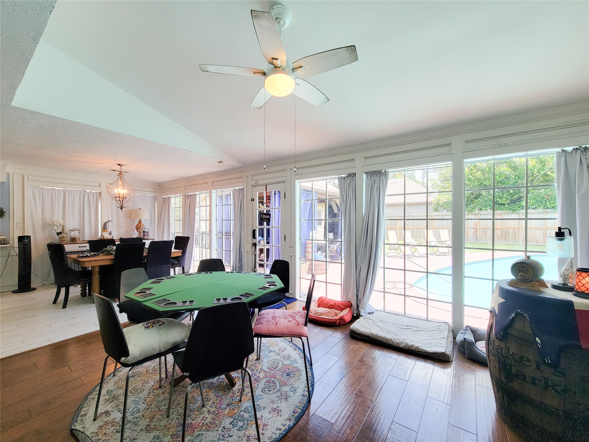 This inviting room features a spacious open layout with large windows overlooking a backyard pool. It includes a dining area with elegant lighting and a cozy seating space, ideal for entertaining and relaxation.