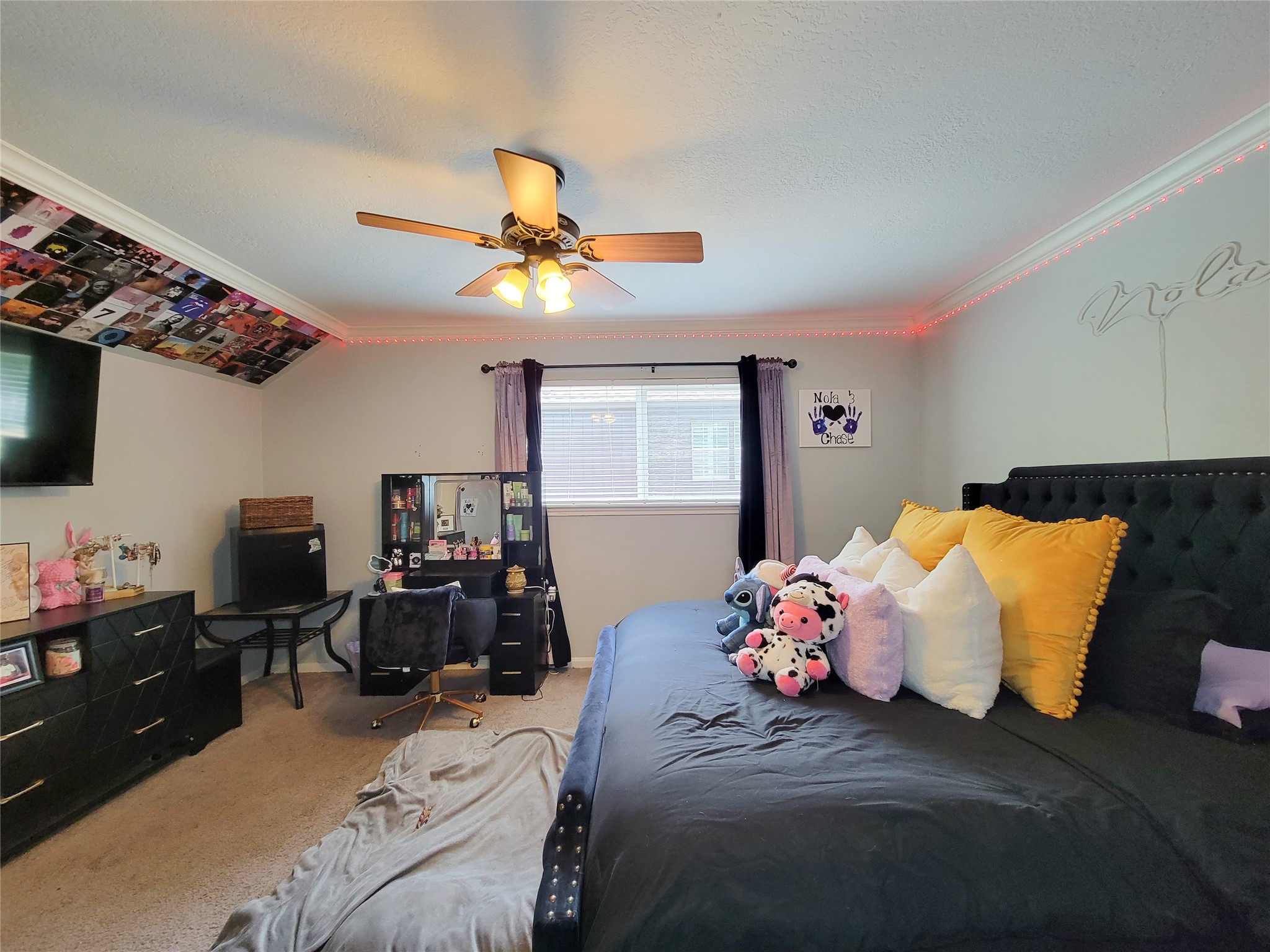 This cozy bedroom features soft carpeting, a modern ceiling fan, and ample natural light from a large window. It includes a stylish bed setup with vibrant pillows, a workspace with a desk and chair, and a dresser for storage. Decorative lighting and personal touches add charm to the space.