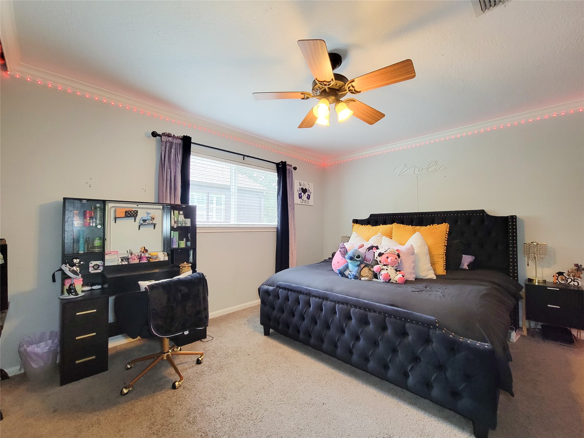 Bright bedroom with a large window, ceiling fan, and cozy carpet. Features a stylish black bed with plush pillows and a charming vanity desk. Ideal for comfort and relaxation.