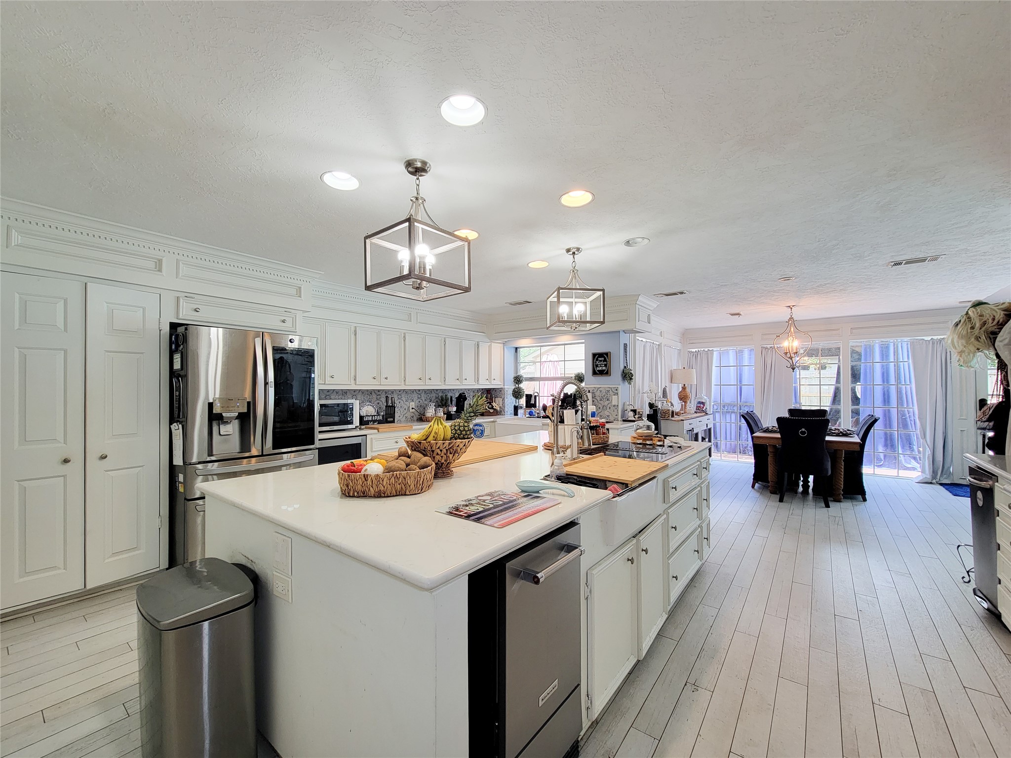 This kitchen features a spacious, open layout with modern stainless steel appliances and a large central island. It has ample white cabinetry, hardwood floors, and recessed lighting. The adjoining dining area is bright, thanks to large windows and glass doors leading outside.
