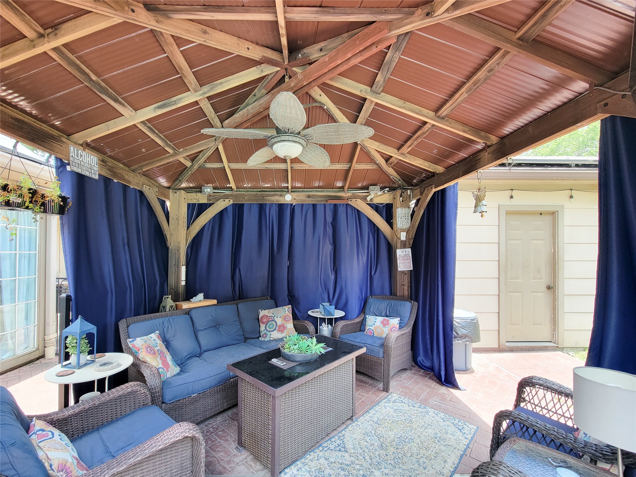 Cozy outdoor patio with a wooden gazebo, featuring comfortable wicker seating, a ceiling fan, and blue curtains for privacy. It's an inviting space for relaxation and entertaining.