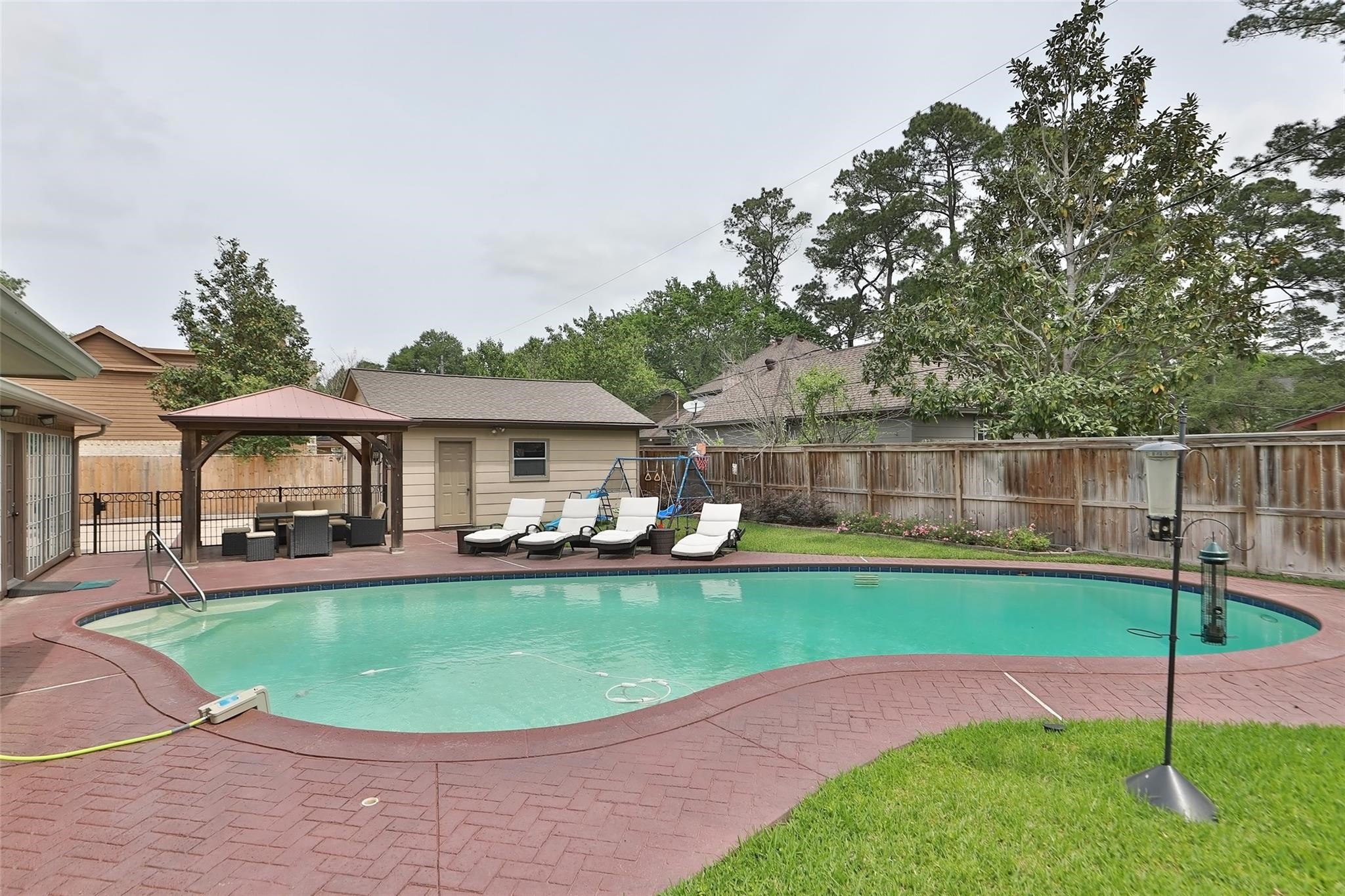 This backyard features a spacious, curved swimming pool surrounded by a brick patio. The area is enclosed by a wooden fence and bordered by mature trees, offering privacy and a serene atmosphere. This backyard features a spacious, curved swimming pool surrounded by a brick patio. The area is enclosed by a wooden fence and bordered by mature trees, offering privacy and a serene atmosphere.