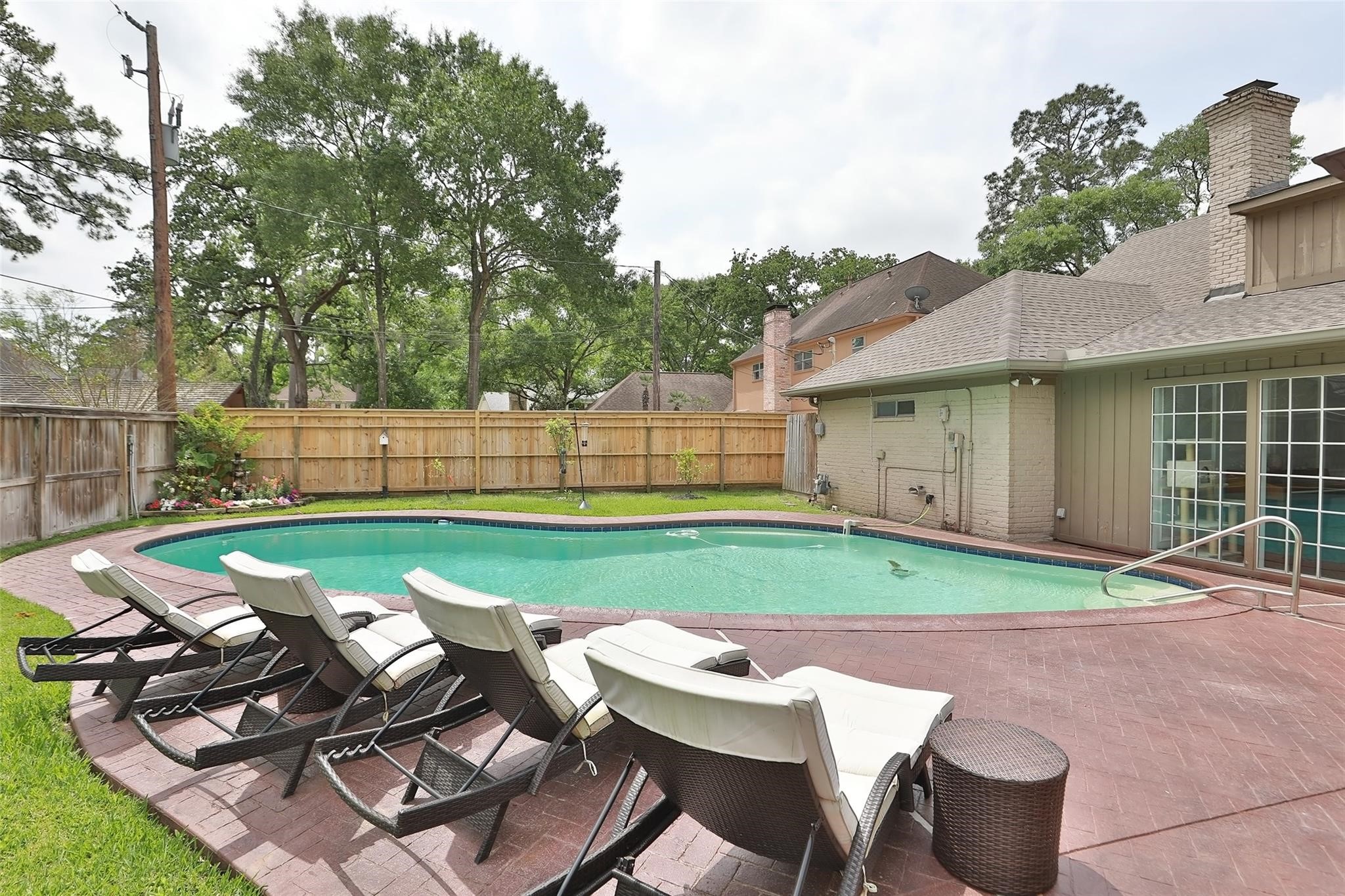 Photo showcases a backyard with a swimming pool surrounded by a brick patio. There are several lounge chairs for relaxation and the area is enclosed by a wooden fence, providing privacy. Mature trees in the background offer shade and a serene atmosphere.