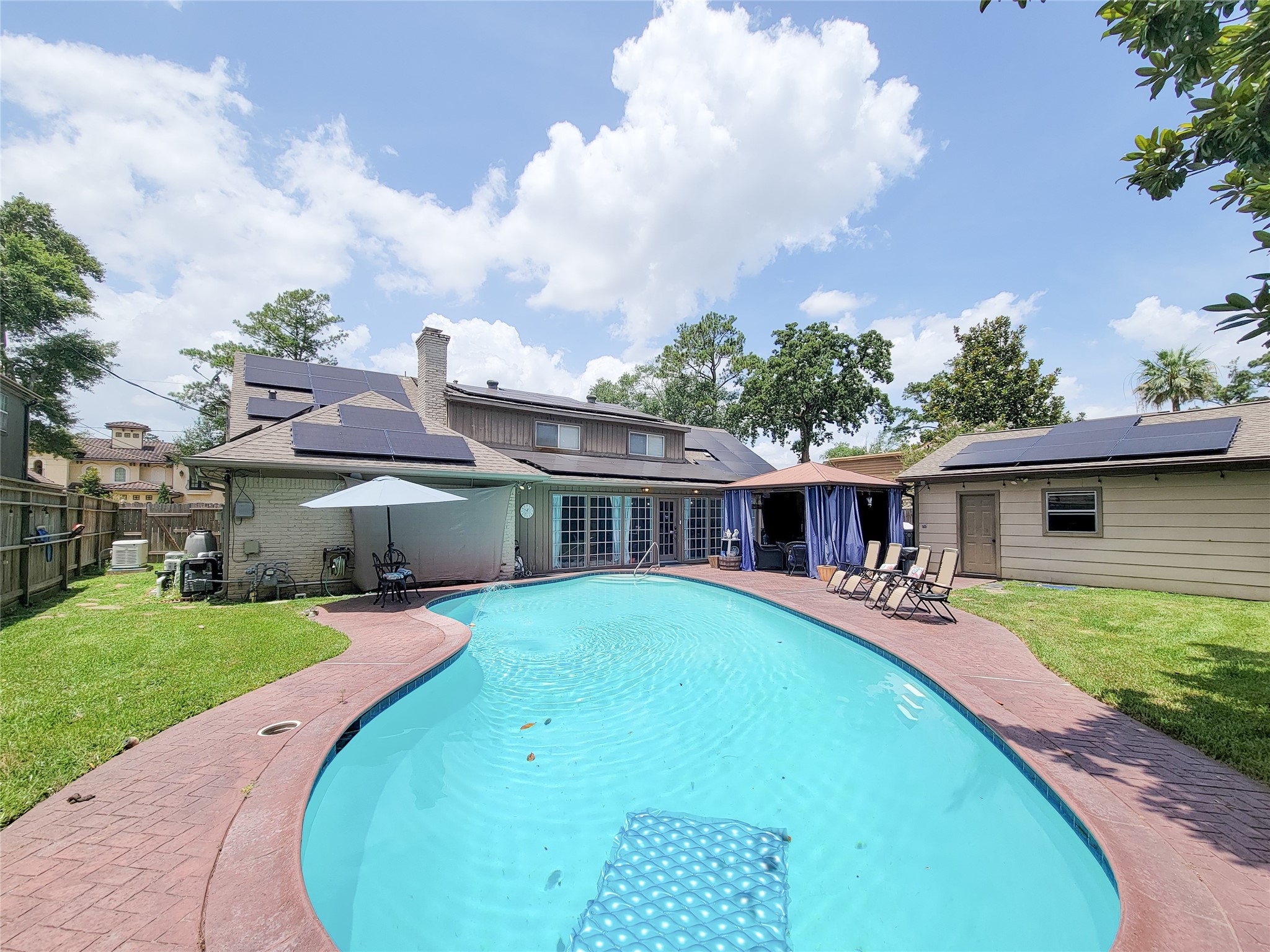 This backyard features a spacious swimming pool with a curved design, surrounded by a paved deck. There's a cozy seating area with an umbrella and a shaded patio with curtains for privacy. Solar panels are installed on the roof, and the area is fenced for added security. Lush greenery completes the serene setting.