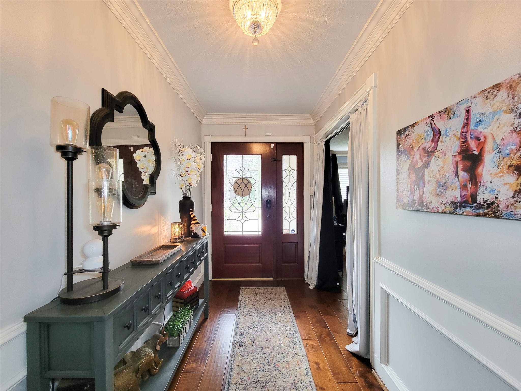This welcoming entryway features a wooden front door with decorative glass panels, elegant wainscoting, and hardwood flooring. A stylish console table with mirror and art accents adds charm, complemented by soft lighting from a ceiling fixture.