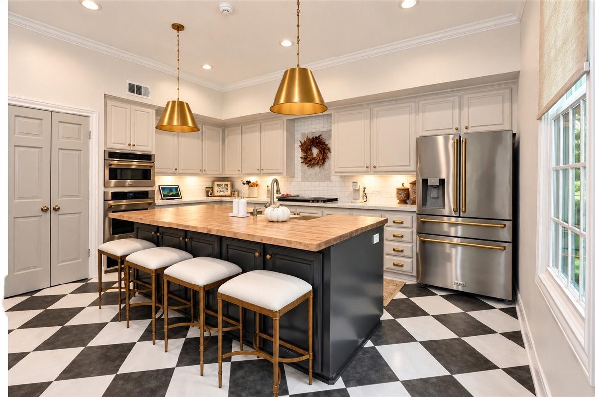 This refined kitchen pairs a wood-block island with seating for four and storage below, beautiful pendant lights overhead, and new Z Line stainless and champagne-gold appliances, all against an updated black-and-white tile floor.