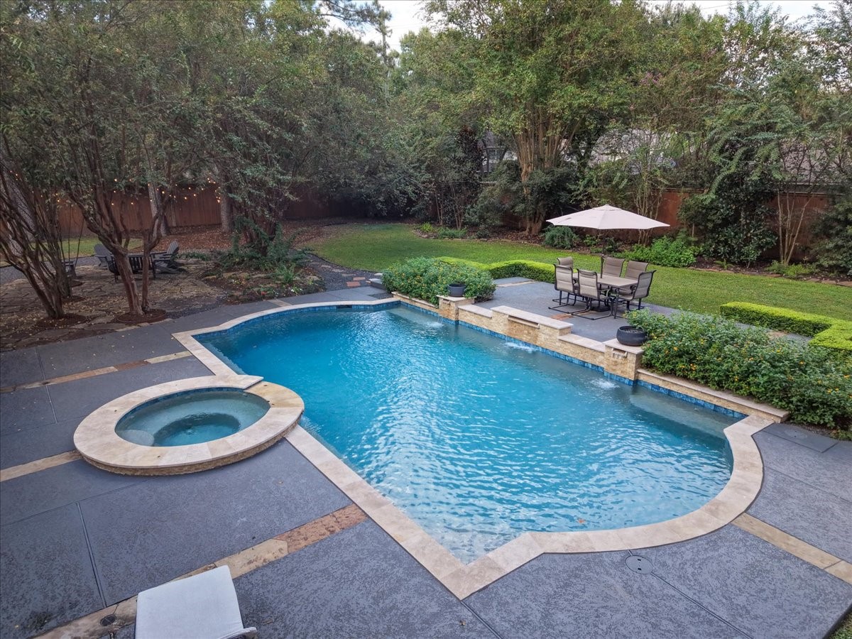 A raised concrete seating area beside the pool, framed by manicured hedges, is made for outdoor dining and lounging, with mature crape myrtles and trees for shade and privacy.