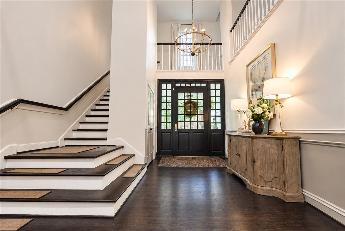 Step into a grand first impression in this dramatic two-story foyer, where a sweeping staircase with dark treads and crisp white risers, classic wainscoting, and a gleaming chandelier create an inviting arrival.