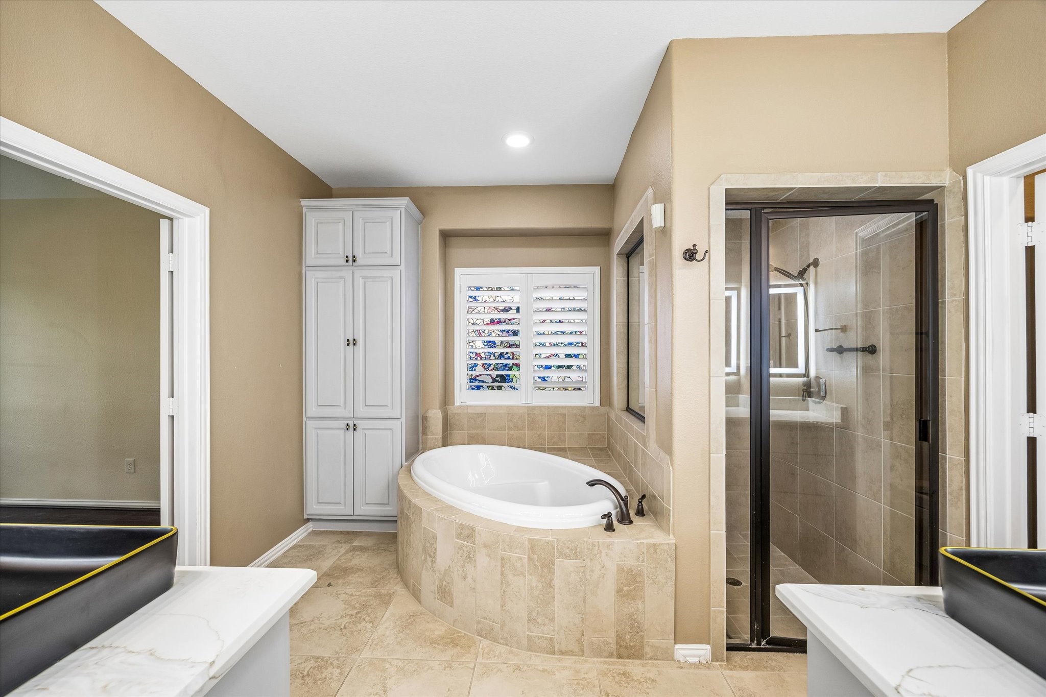 The primary en-suite bath offers separate vanities with vessel sinks, a soaking tub beneath a shuttered window and a glass-enclosed shower.  A tall linen cabinet provides ample storage.