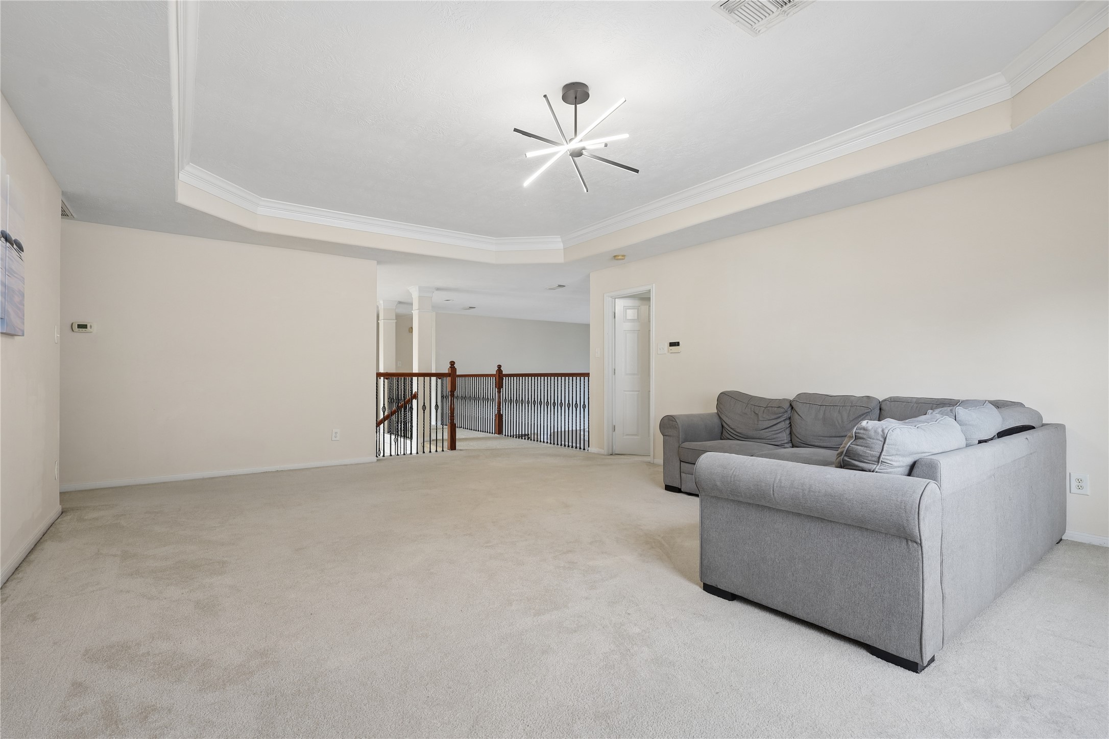 Spacious, carpeted upstairs living area with neutral tones, featuring a modern ceiling light fixture and a cozy gray sectional sofa. The room opens to a railing overlooking the staircase, adding an airy feel. Ideal for relaxation or entertaining.