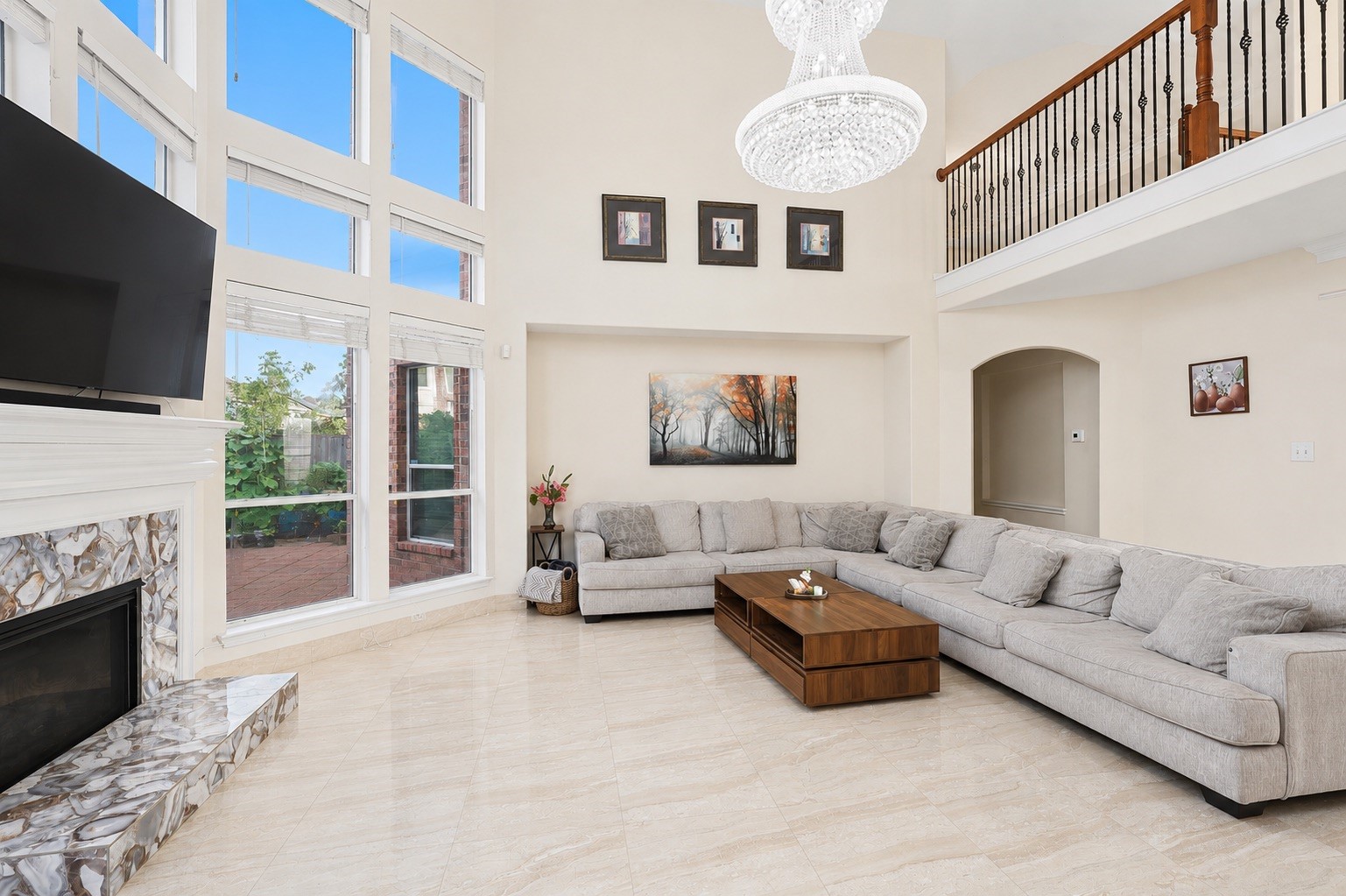 This spacious living room features high ceilings, large windows for natural light, a modern chandelier, and a cozy sectional sofa. It includes a stylish fireplace, elegant tile flooring, and a wooden coffee table, creating a welcoming and luxurious atmosphere.