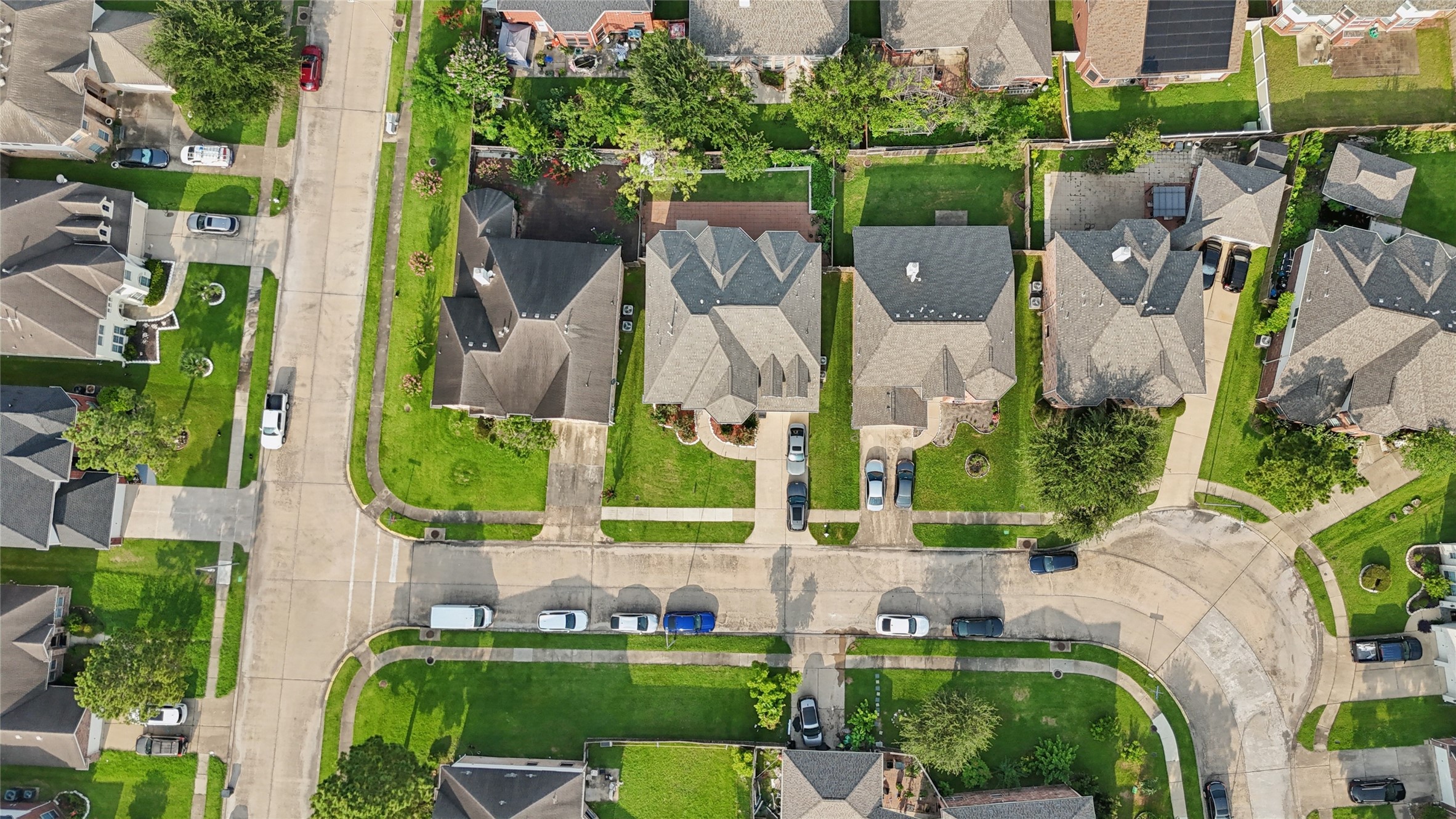 This aerial view showcases a suburban neighborhood with well-maintained homes, neatly landscaped yards, and a curving street layout. The area appears peaceful and residential, with ample parking and green spaces.