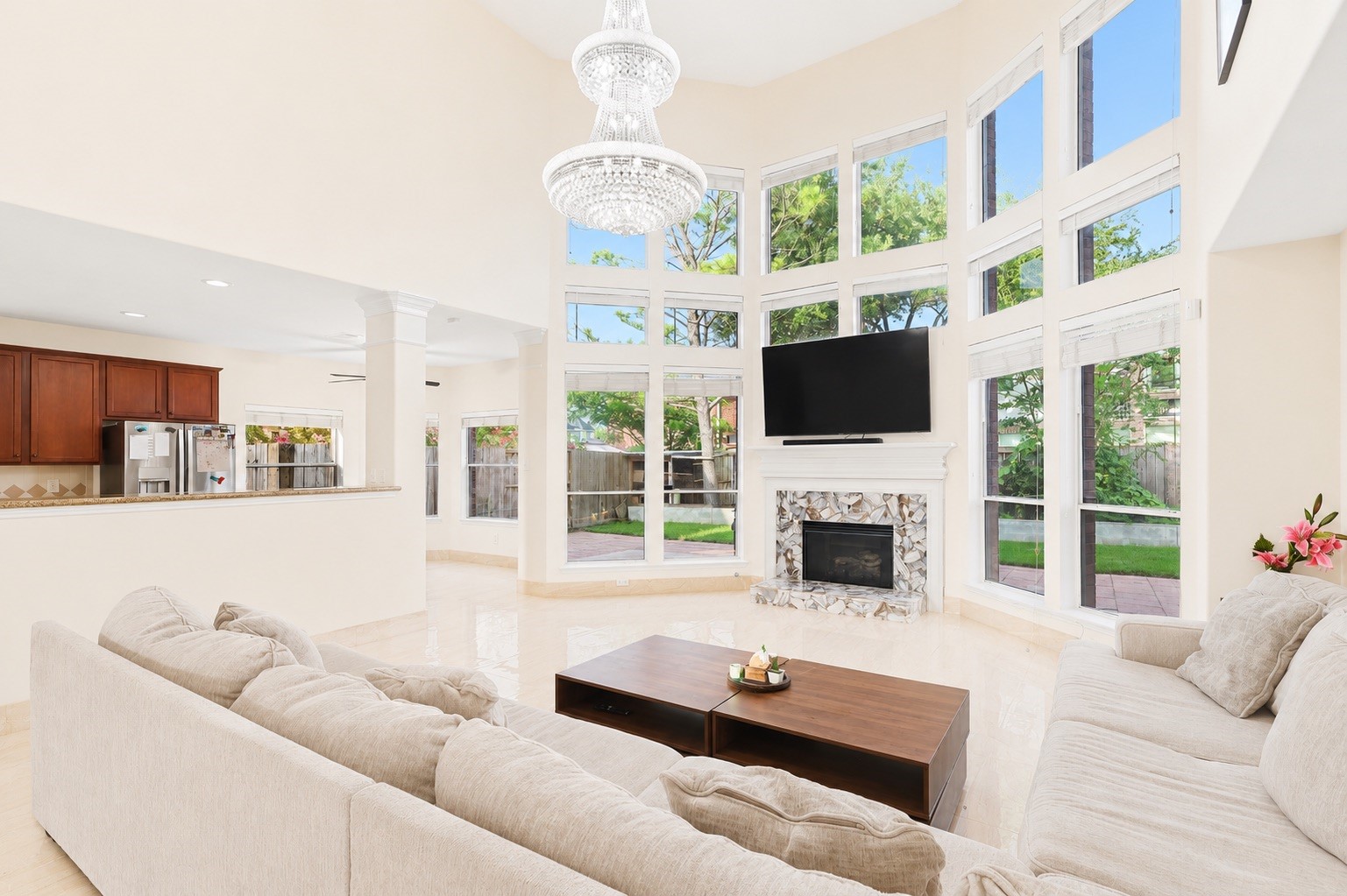 This spacious living room features high ceilings, abundant natural light from large windows, and a cozy fireplace. The open layout flows into the kitchen, offering a modern and welcoming atmosphere perfect for entertaining.