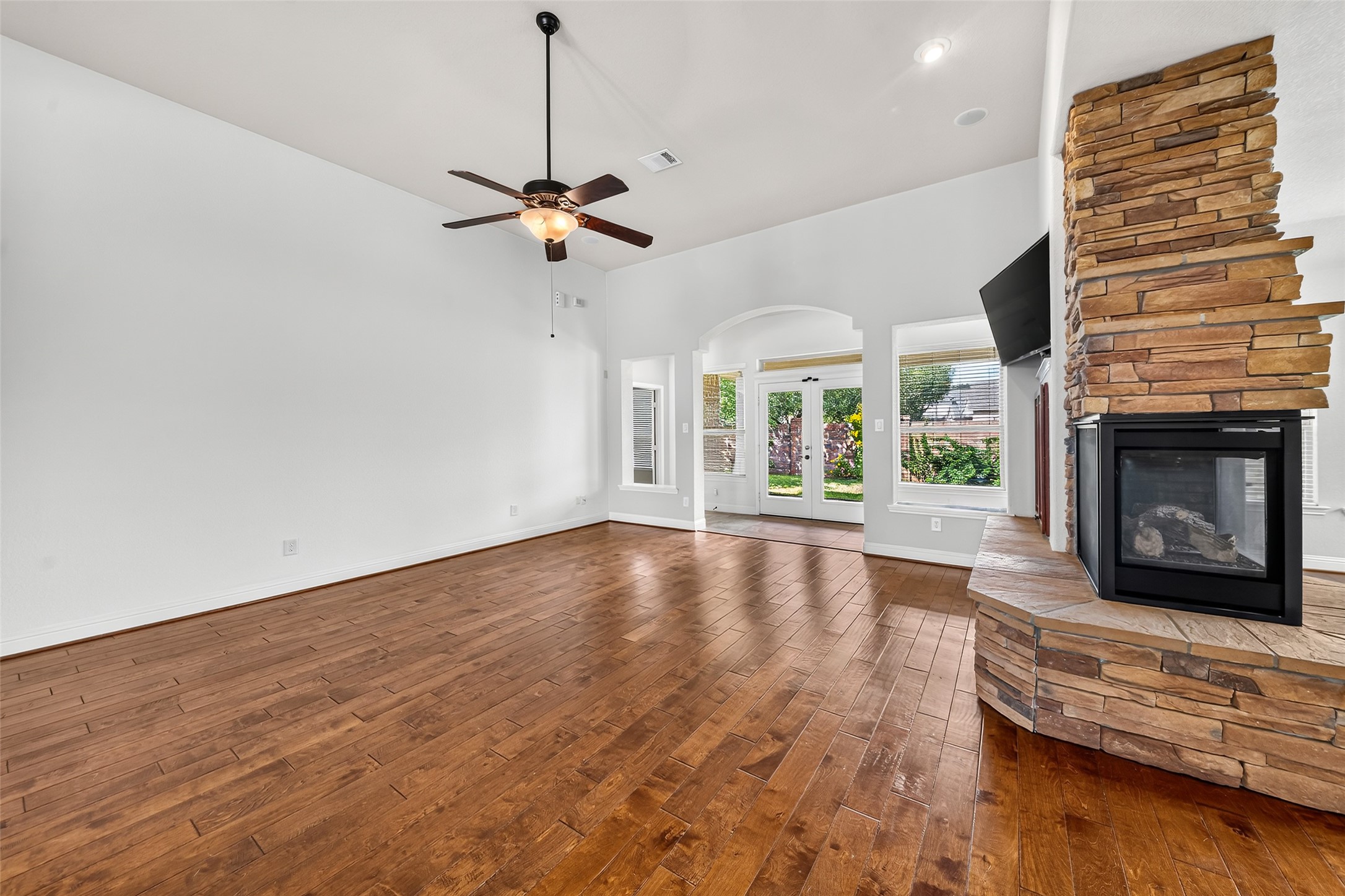 Expansive living room featuring high, elegant ceilings, stunning wooden floors, a charming stone fireplace, ample natural light, and French doors that lead to the backyard.
