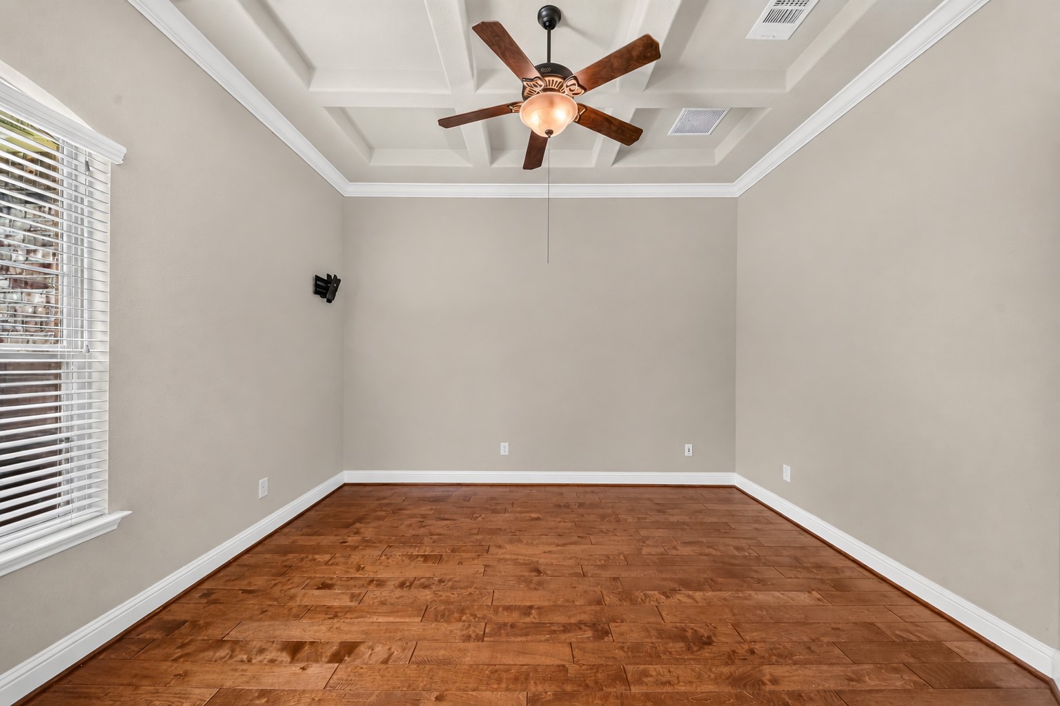 Spacious study featuring elegant wood flooring, a stylish tray ceiling, and natural light from large window.
