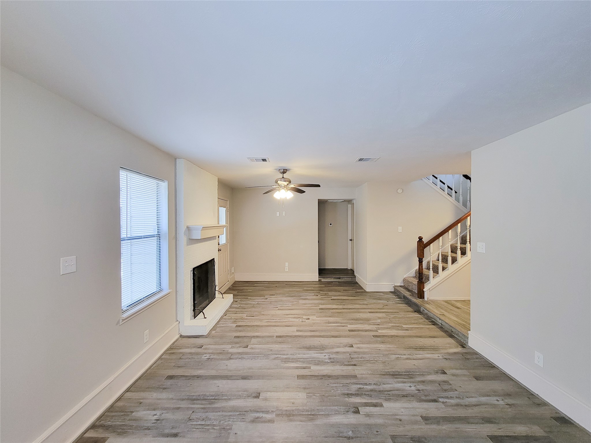 Spacious living area with stylish wood flooring, a cozy fireplace, and a ceiling fan, complemented by natural light from large windows.