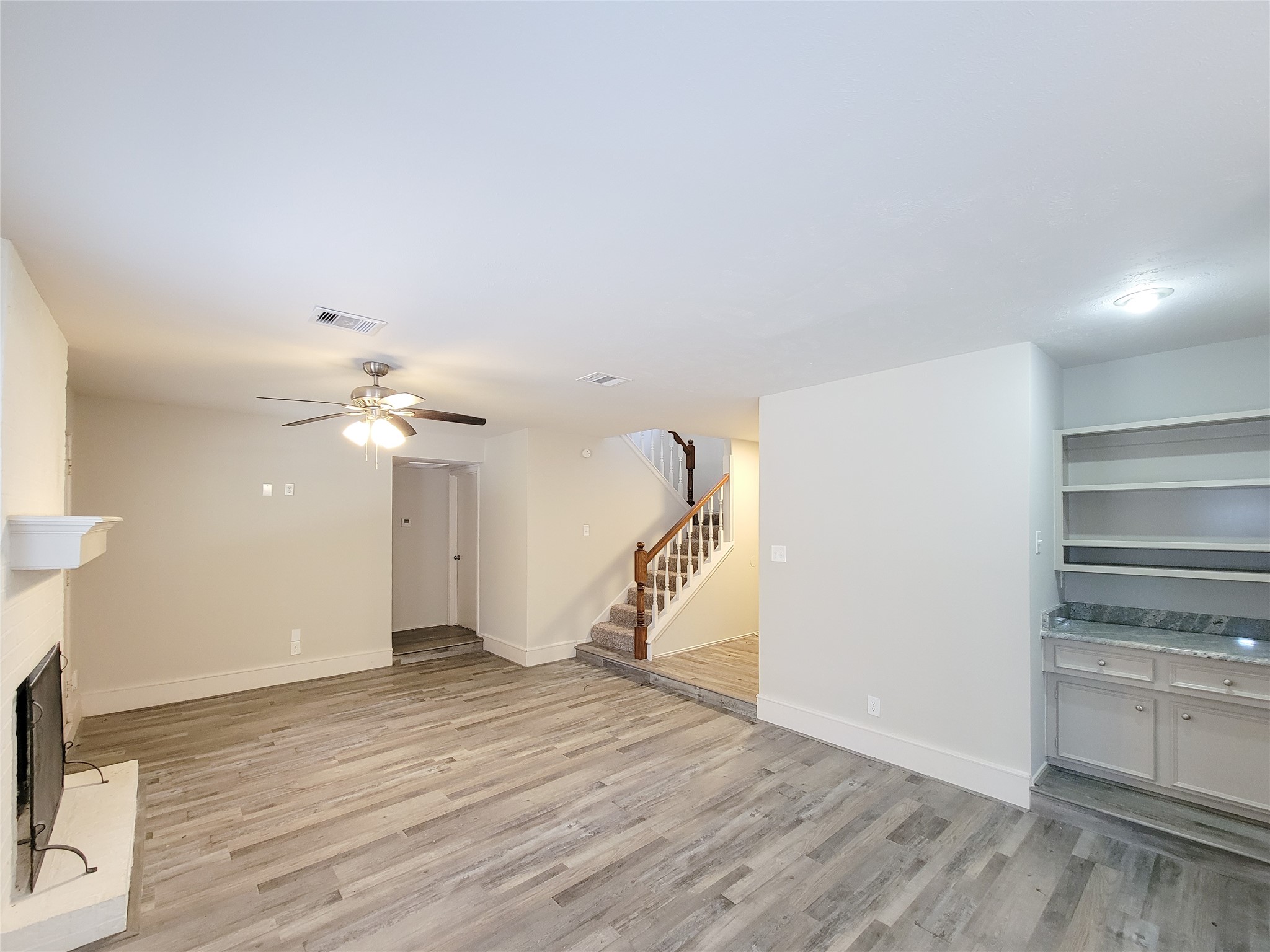 Spacious living area with modern wood-look flooring, a cozy fireplace, built-in shelving, and a ceiling fan, leading to an inviting staircase.