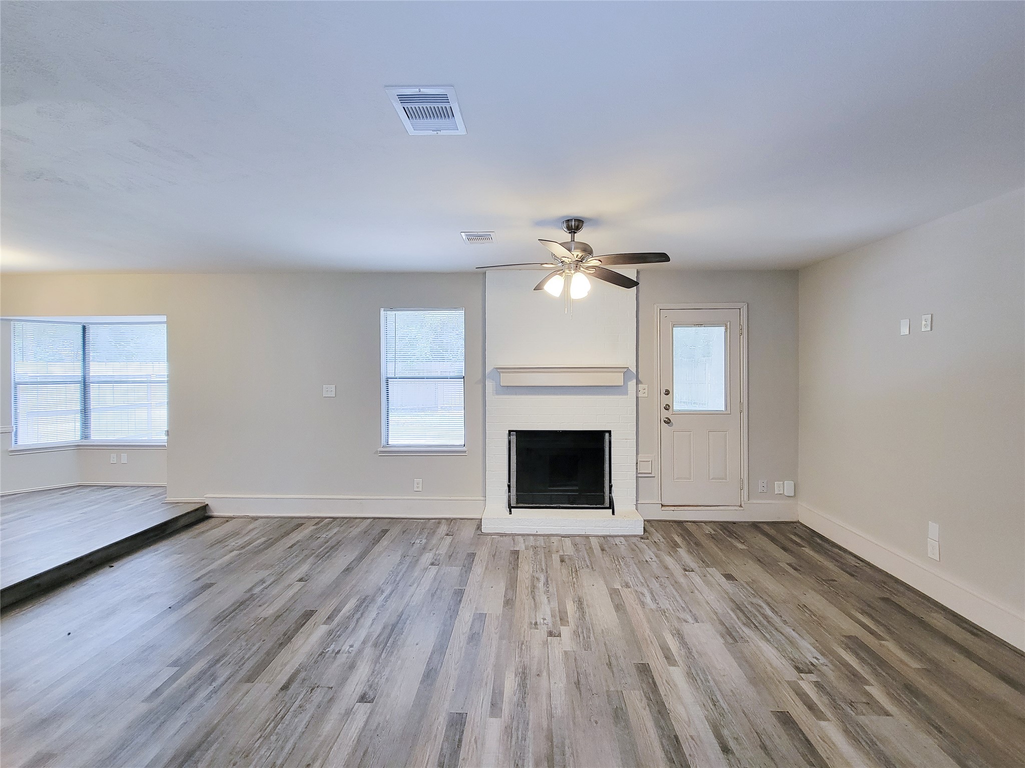 Spacious living room with elegant wood flooring, a cozy fireplace, ceiling fan, and ample natural light through large windows.
