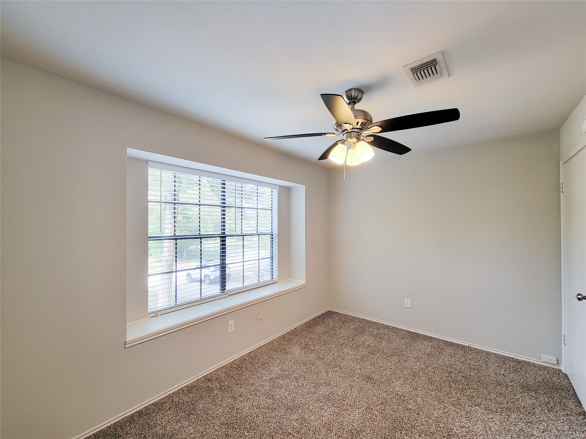 Bright and cozy room featuring a large window with views of greenery, neutral walls, plush carpet, and a stylish ceiling fan.