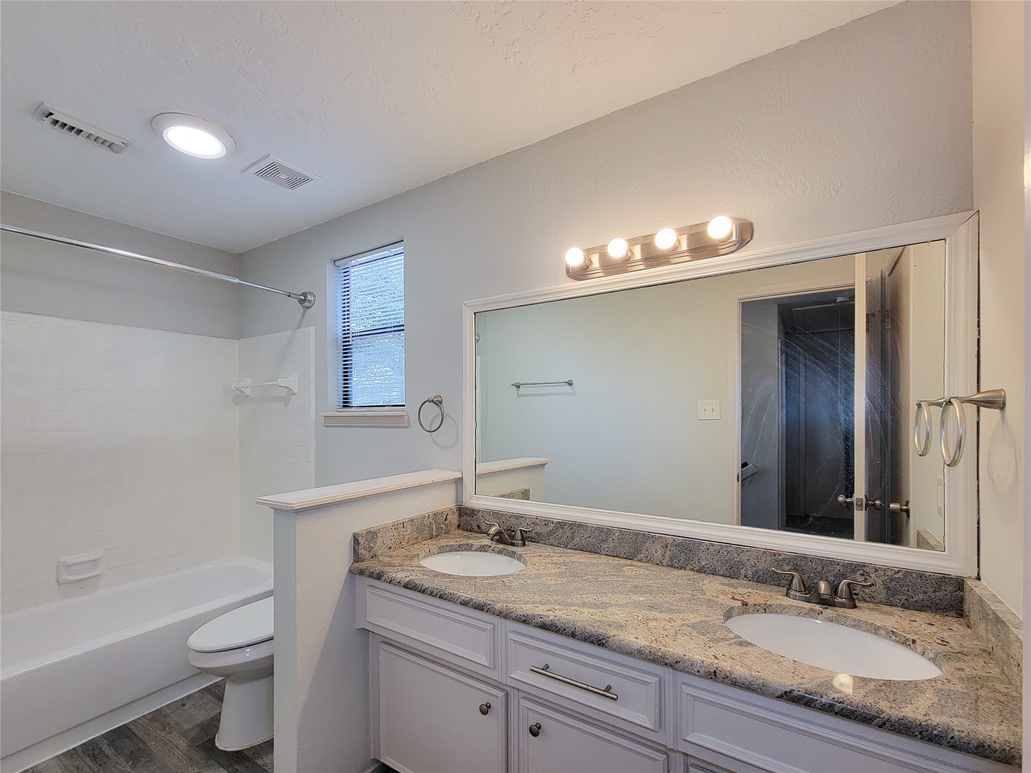 Bright and modern bathroom featuring a double vanity with elegant granite countertops, a spacious mirror, and a classic shower-tub combination.